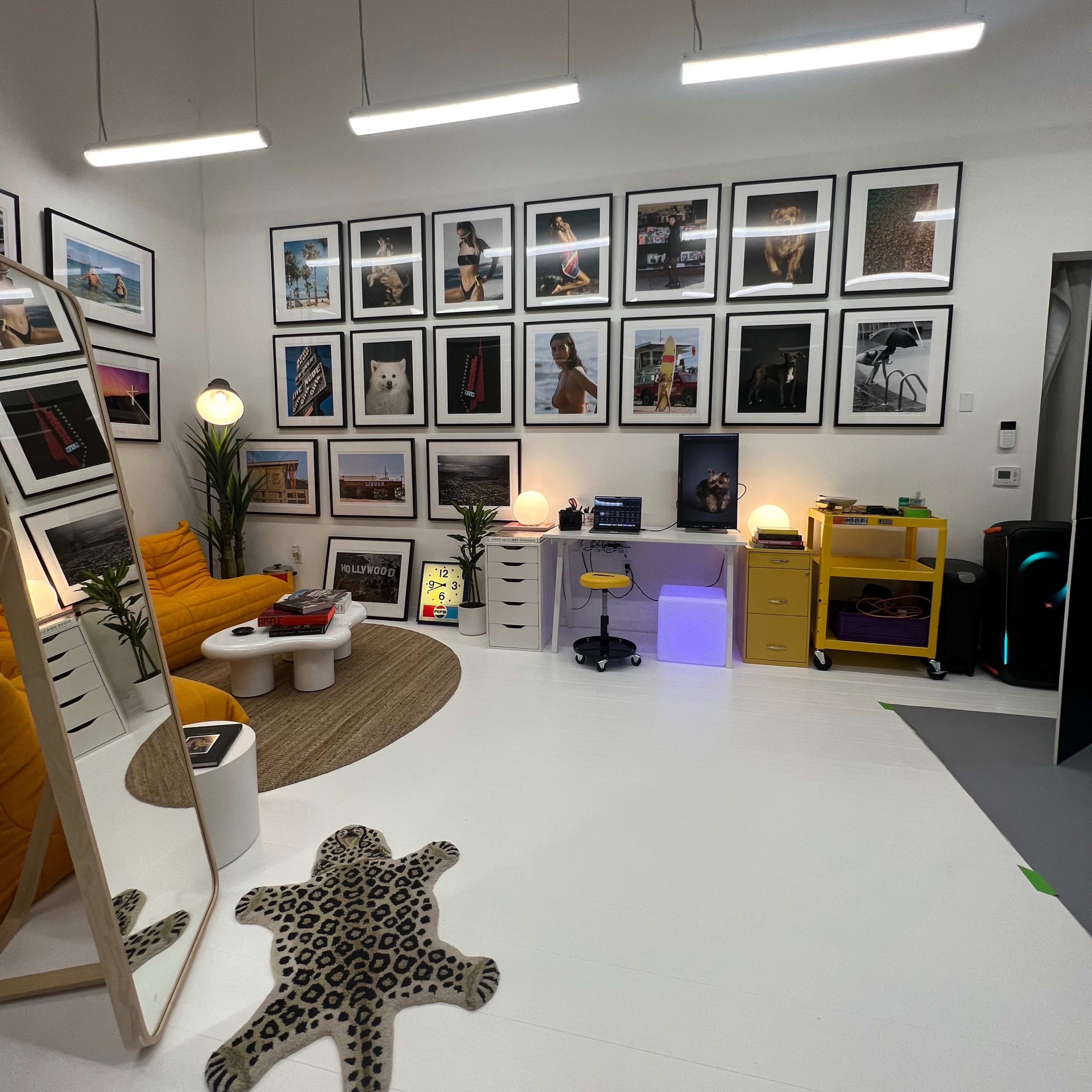 A stylish room featuring a yellow couch, a large mirror, various framed photographs on the wall, and a mix of furniture including a desk and storage units.