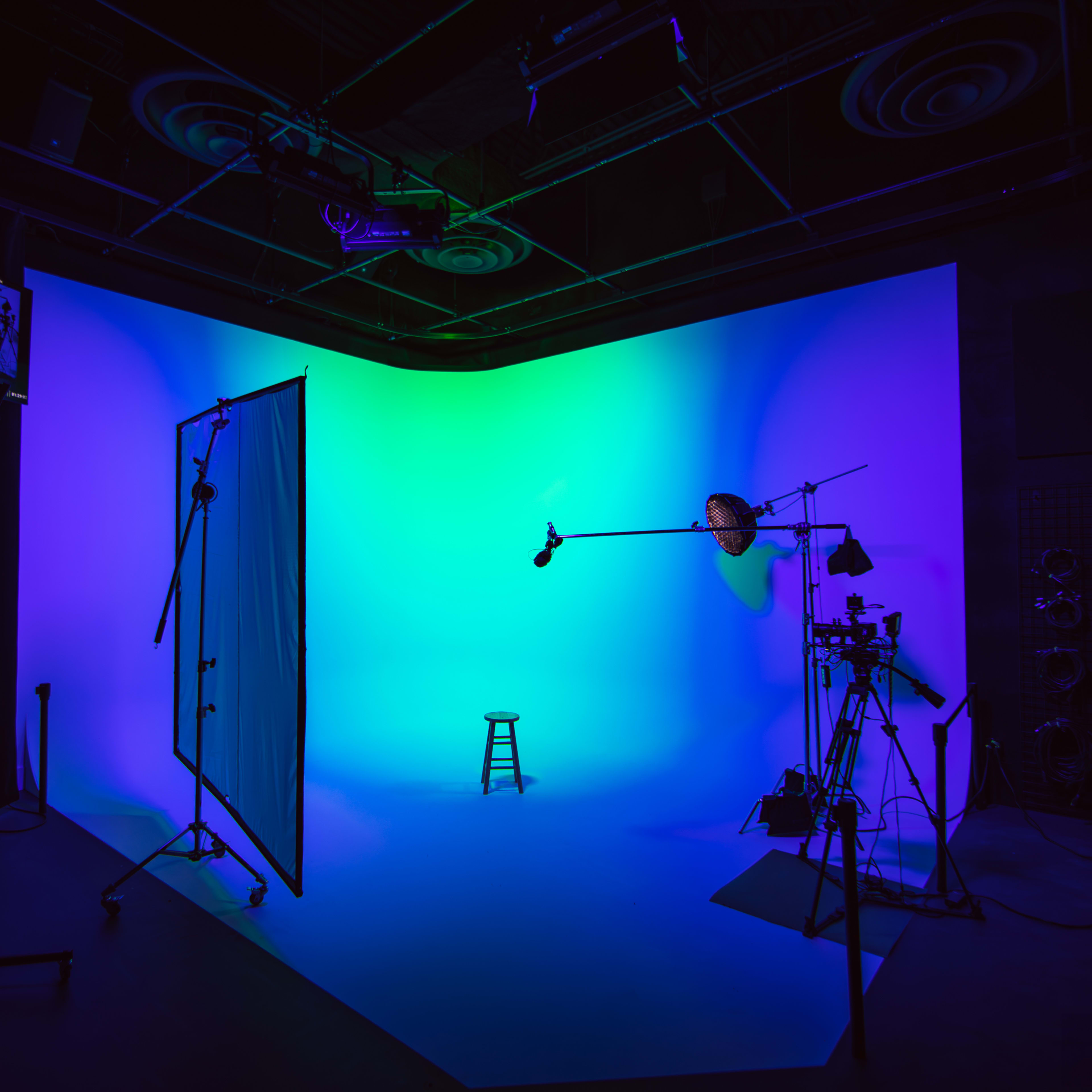 A photography studio with a blue and green backdrop, a stool in the center, and various lighting and camera equipment set up around the area.