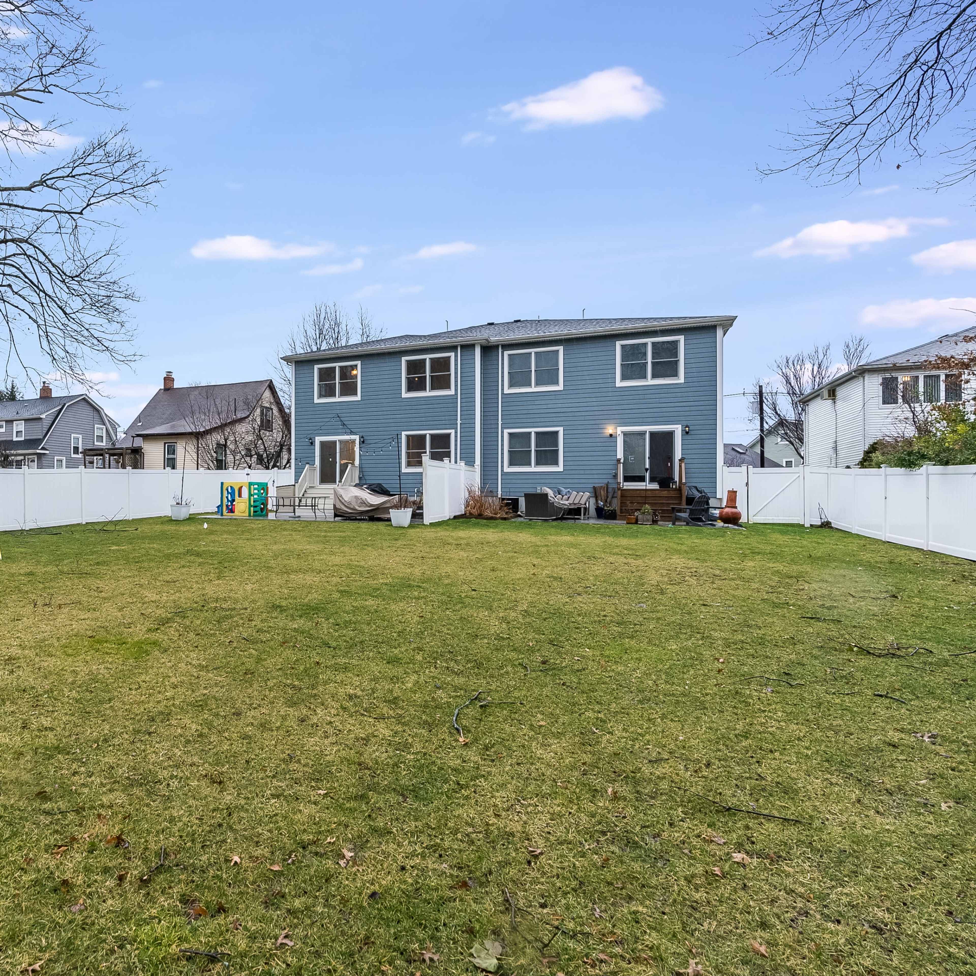 A backyard features a large green lawn with a playset and a blue two-story house surrounded by a white vinyl fence.
