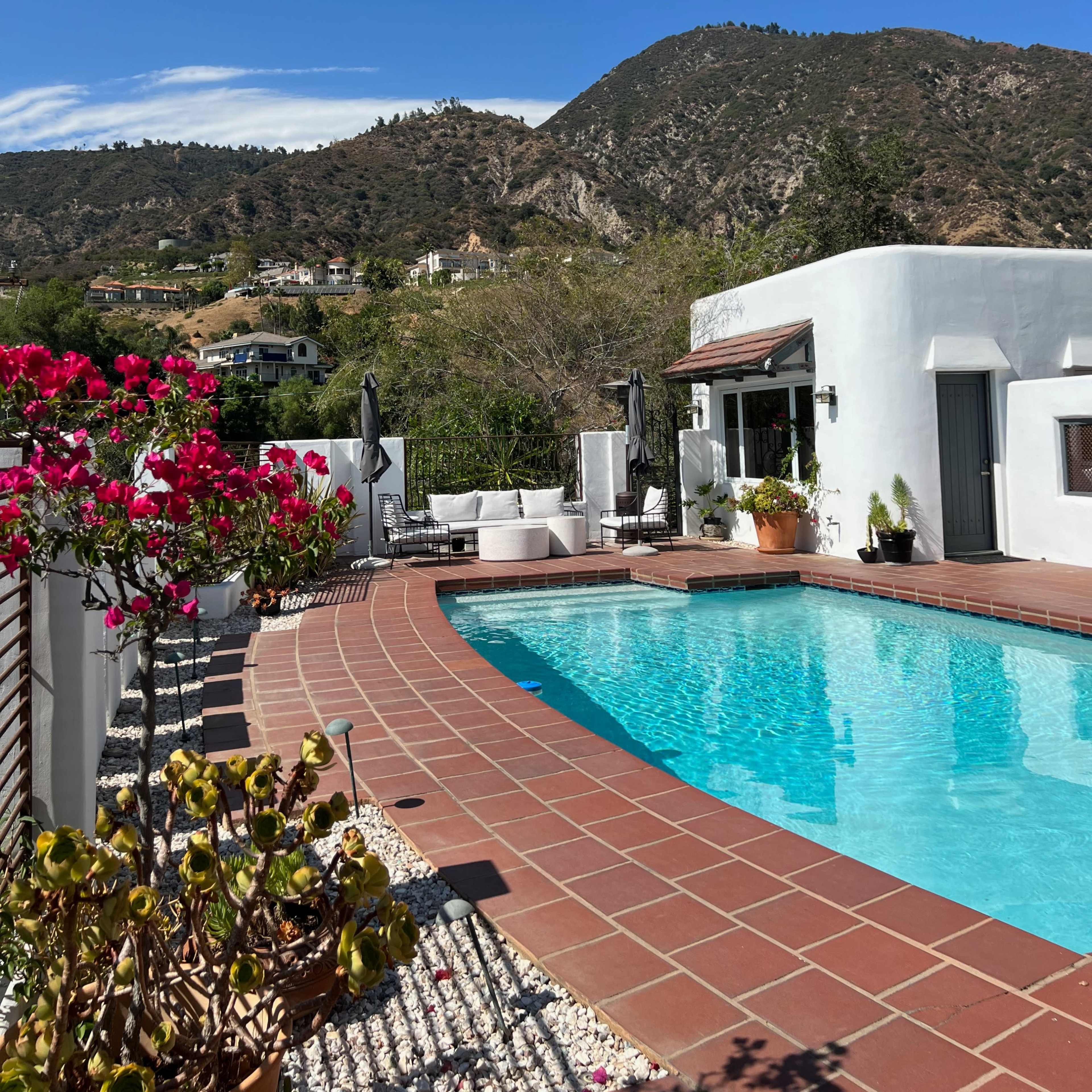 A swimming pool surrounded by a tiled patio, with a backdrop of mountains and blooming flowers.