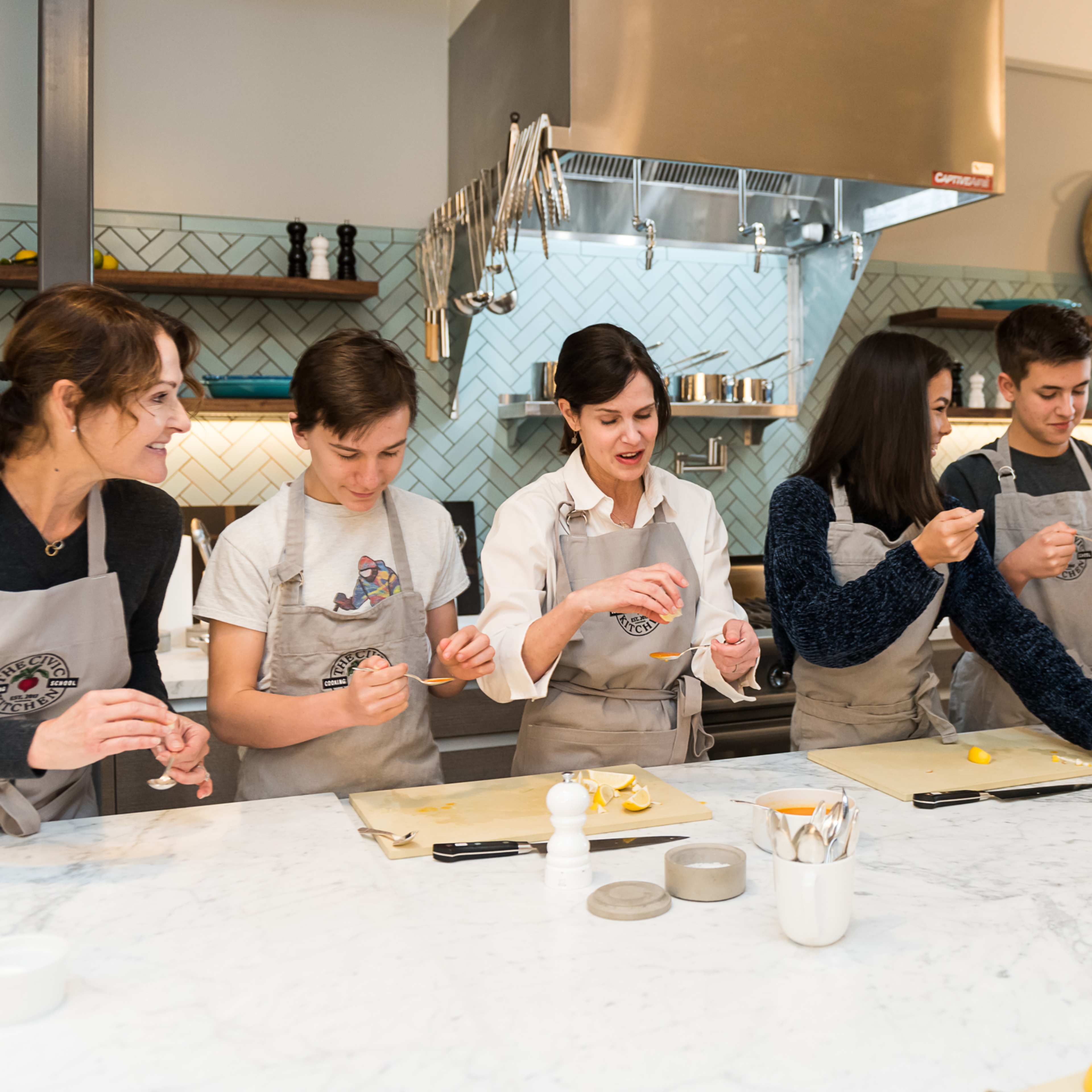 A group of six people is preparing food in a kitchen, each engaged in a cooking task at a marble countertop.