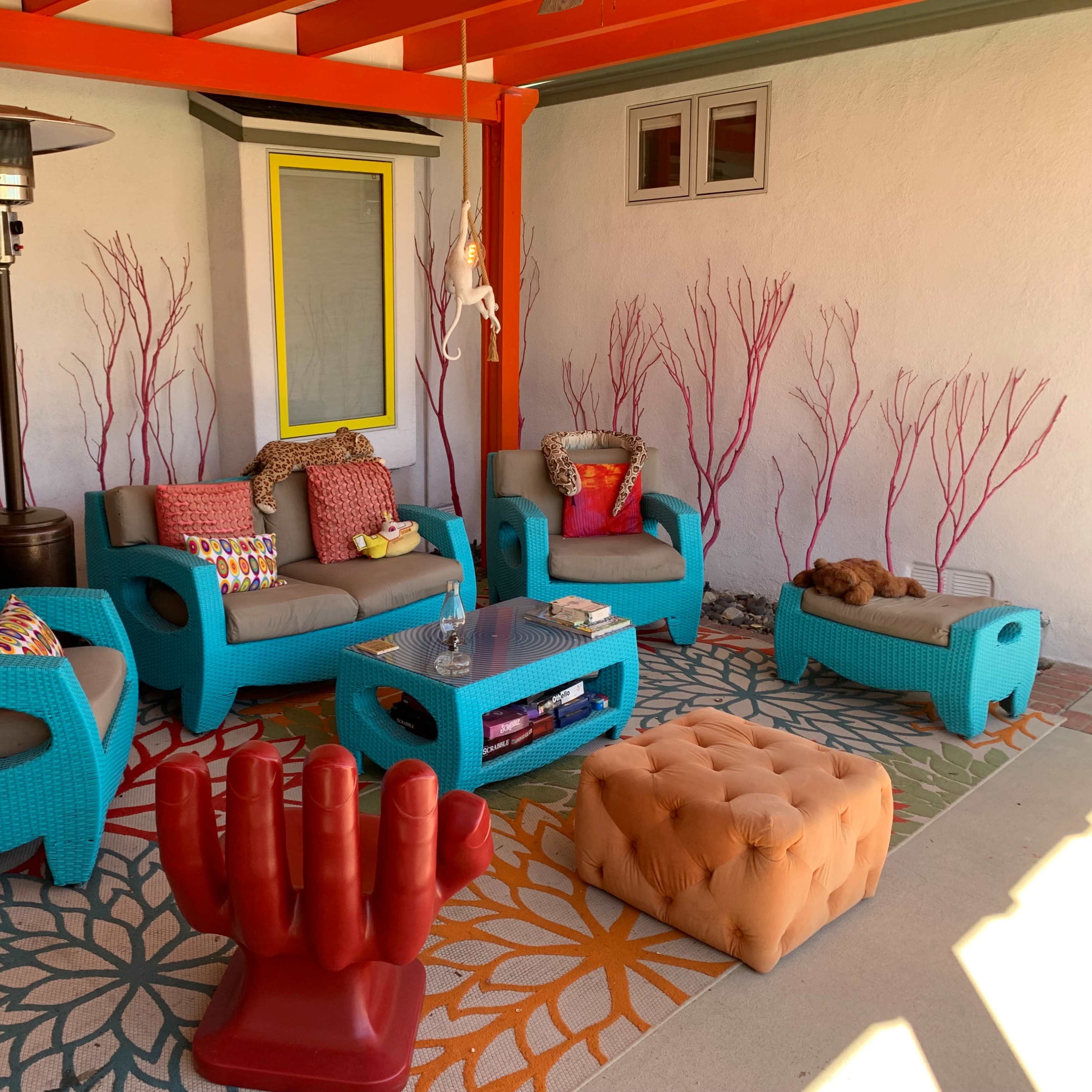 A vibrant outdoor seating area with turquoise furniture, a beige cushioned ottoman, a glass coffee table, and decorative pillows on a colorful patterned rug.