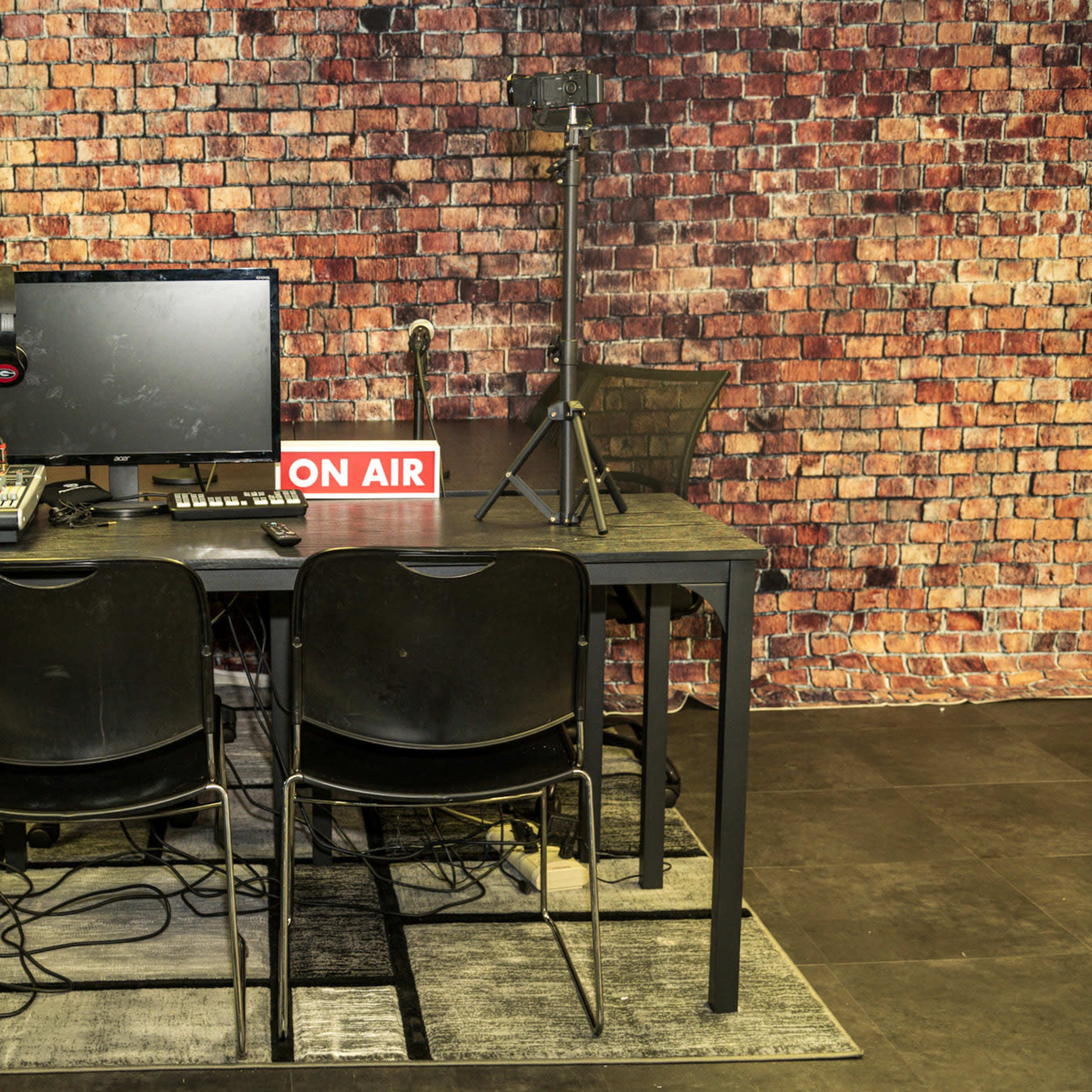 A studio setup features a computer monitor, audio equipment, and two chairs in front of a brick wall, with an "ON AIR" sign visible.