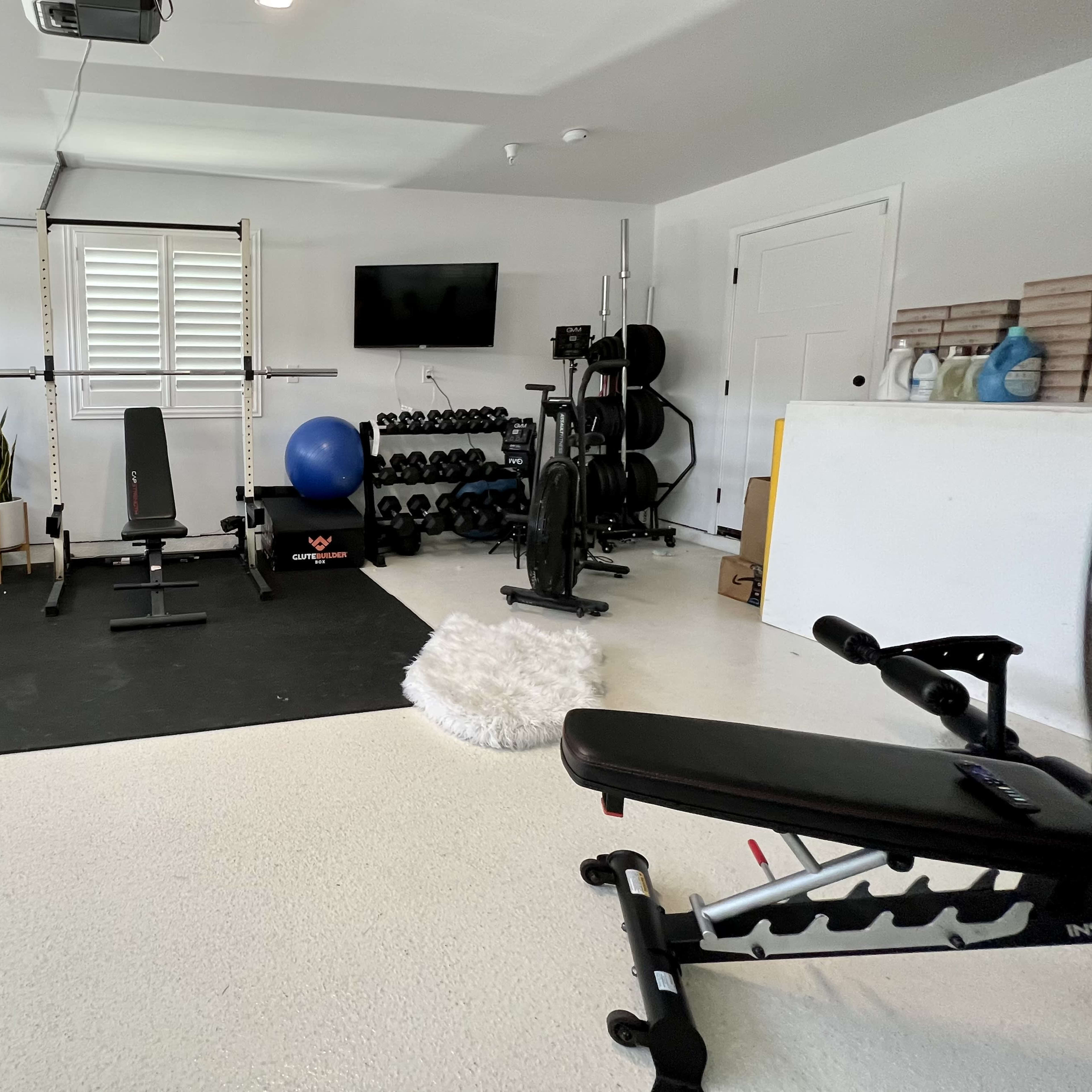 A garage gym setup featuring various exercise equipment, including a treadmill, weight bench, barbell with weights, and a stability ball, all arranged on a rubber mat.