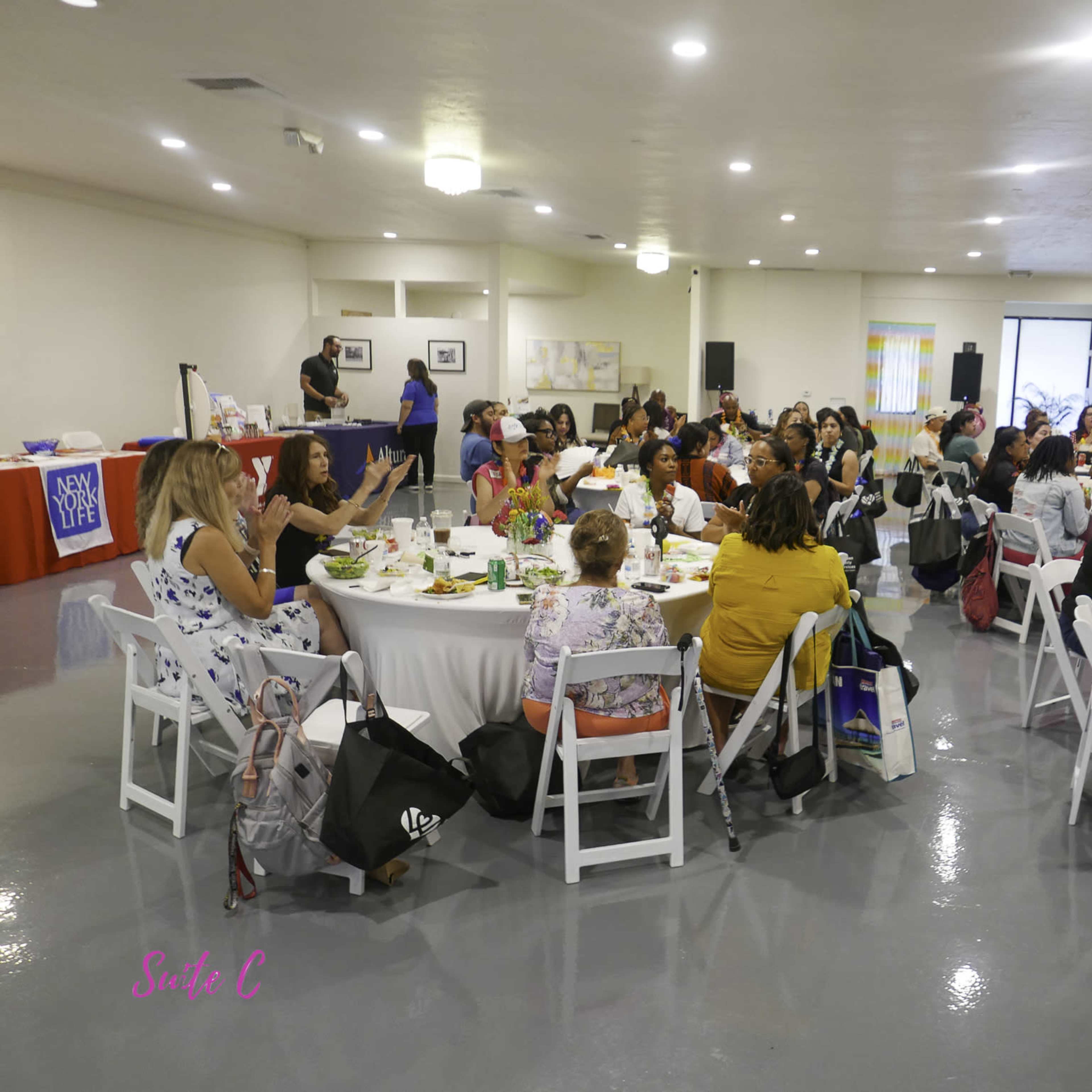 A gathering of diverse women seated at round tables, enjoying food and engaging in conversation in a well-lit event space.