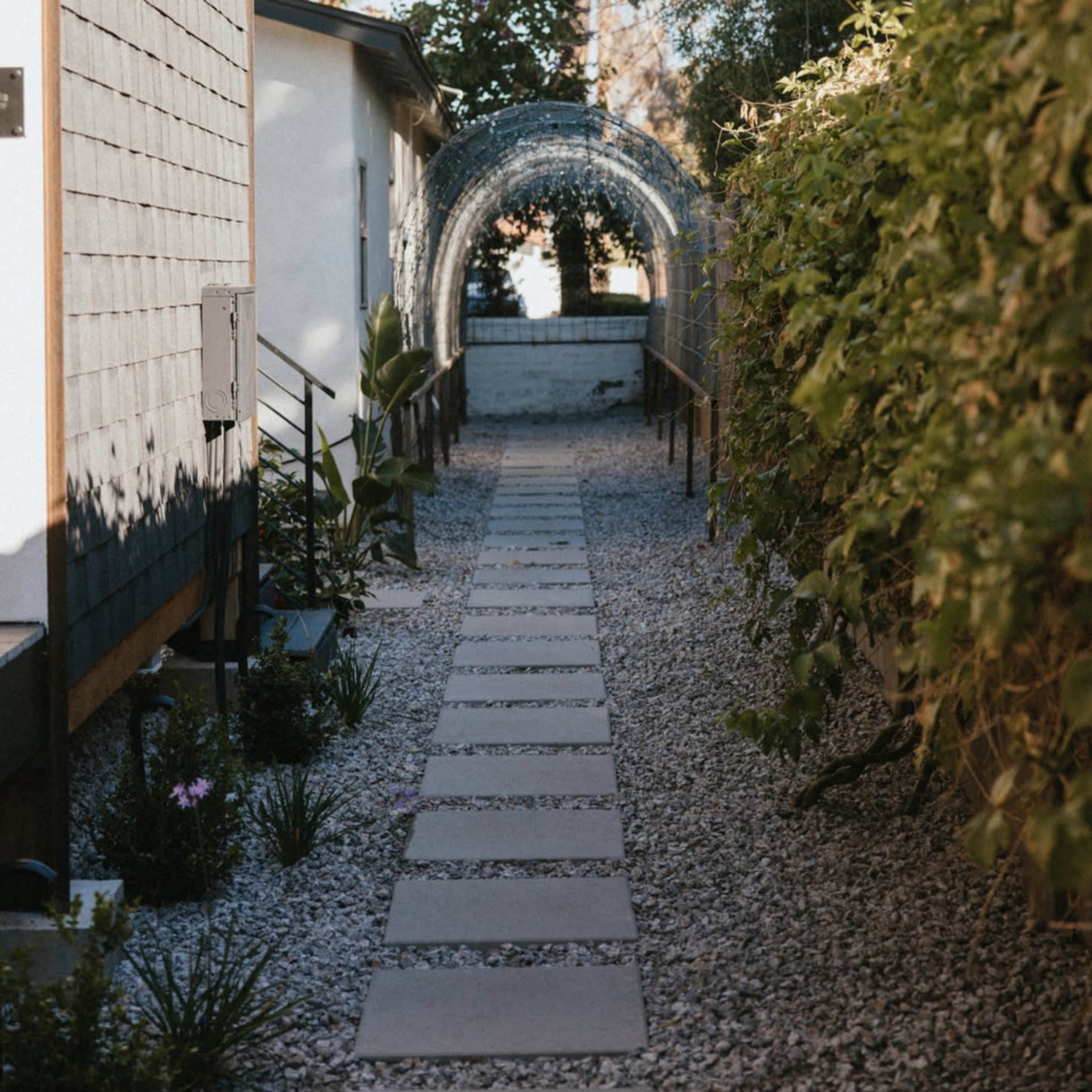 A stone path lined with small plants leads through a trellis-covered archway towards a distant wall.