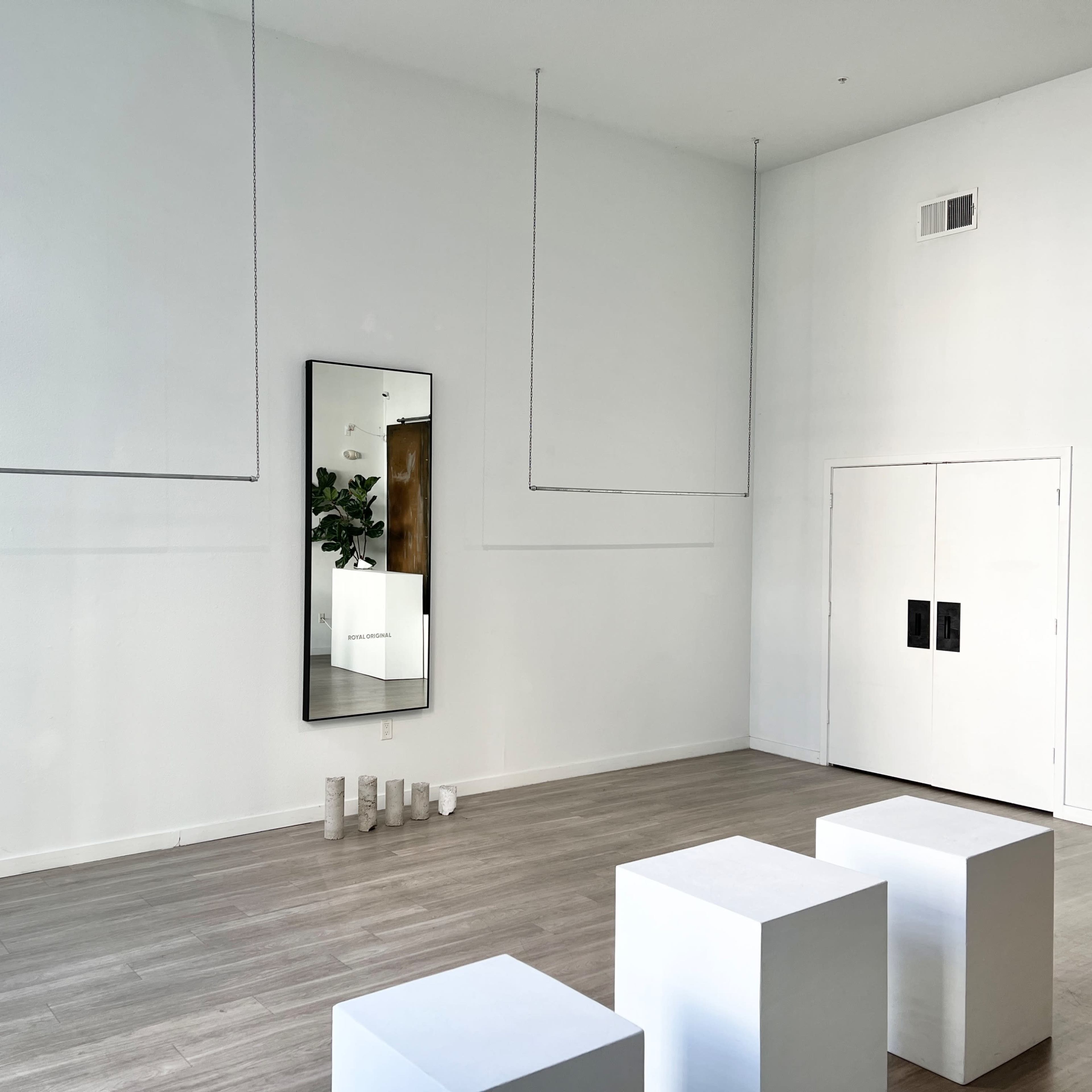 A minimalist interior space with white walls, a large mirror, three white cubes, and a door, along with several candles arranged on the floor.