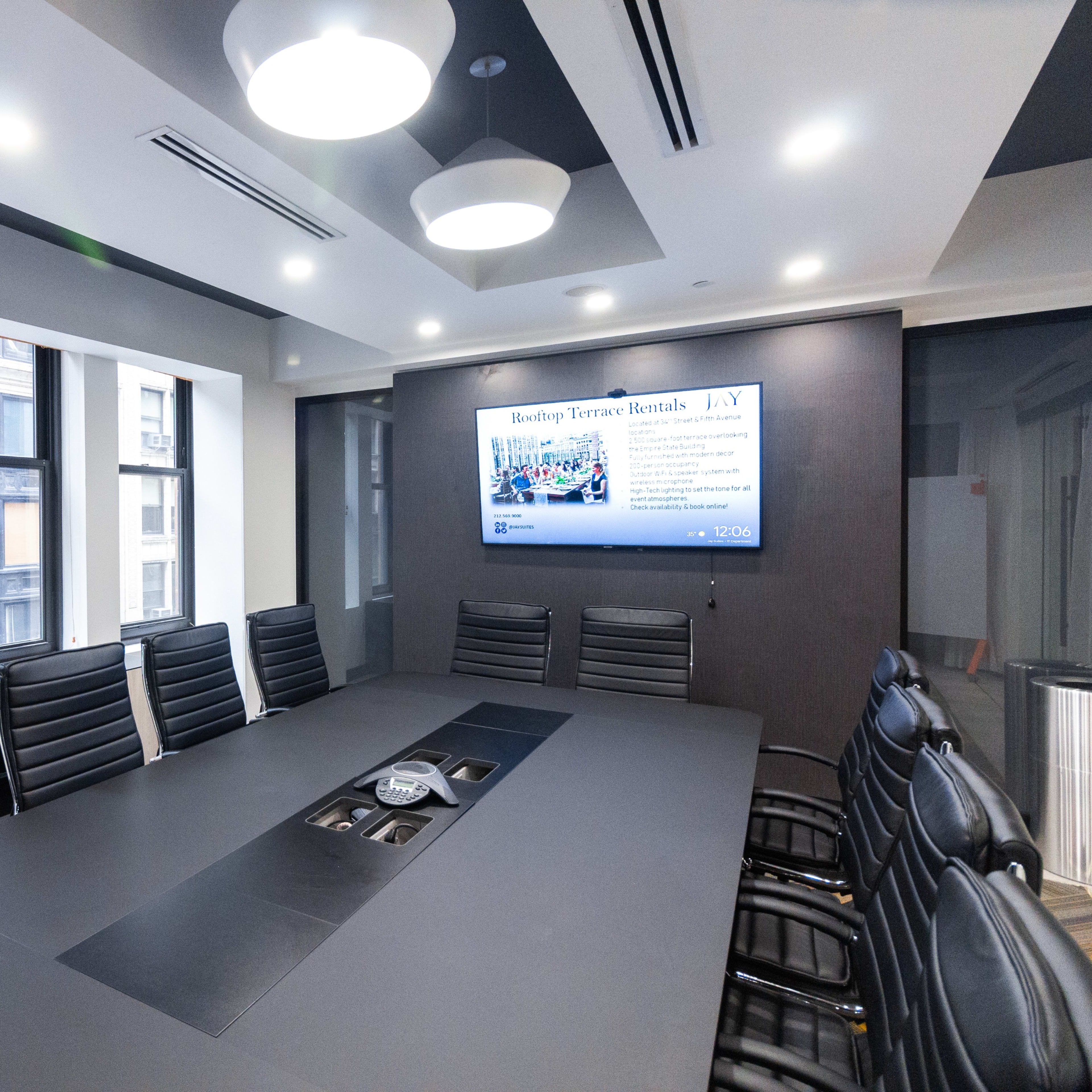 A modern conference room features a large rectangular table surrounded by black leather chairs, with a screen displaying information on one wall and windows letting in natural light.