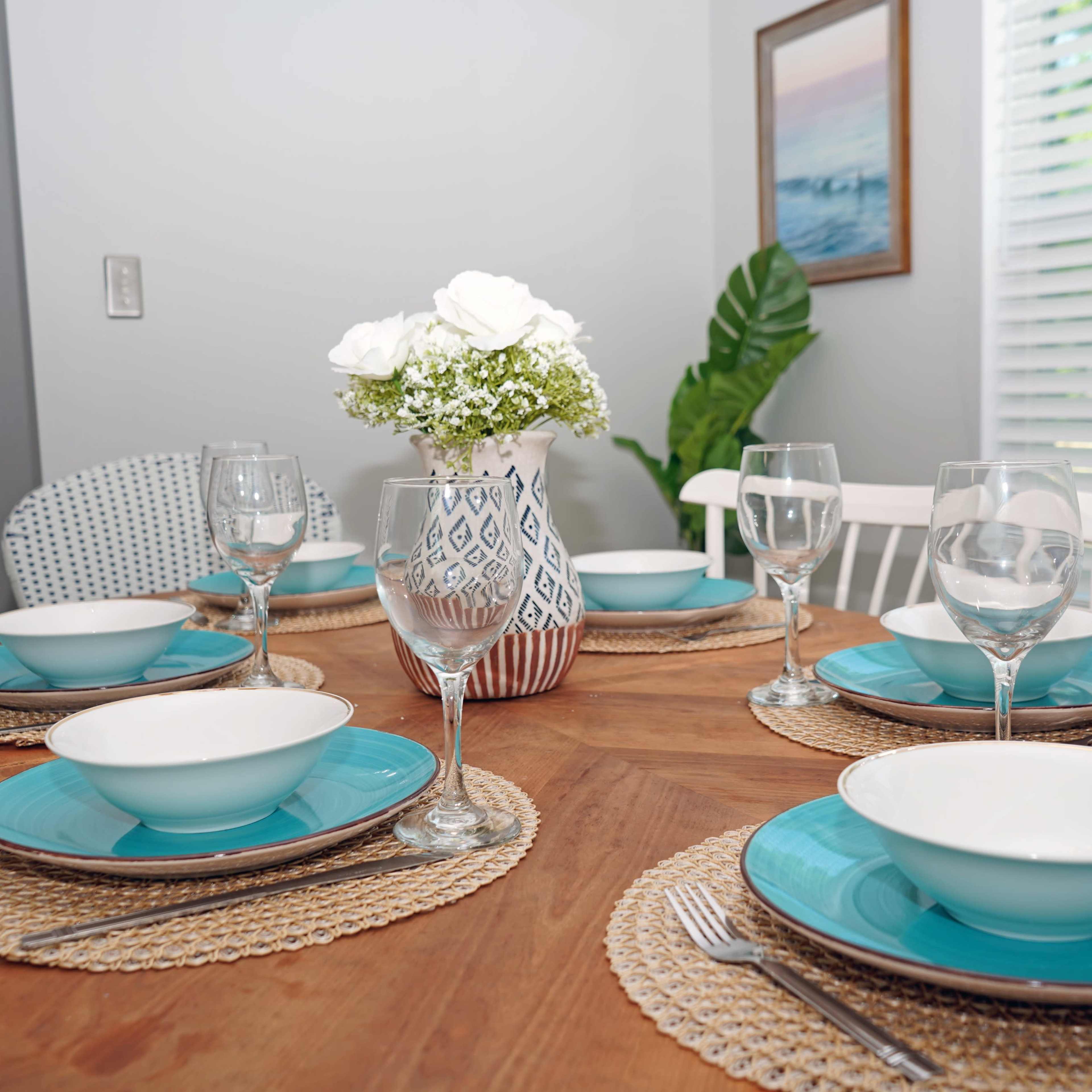 A dining table is set with blue plates, white bowls, and wine glasses, accompanied by a vase of white flowers and surrounded by woven placemats.