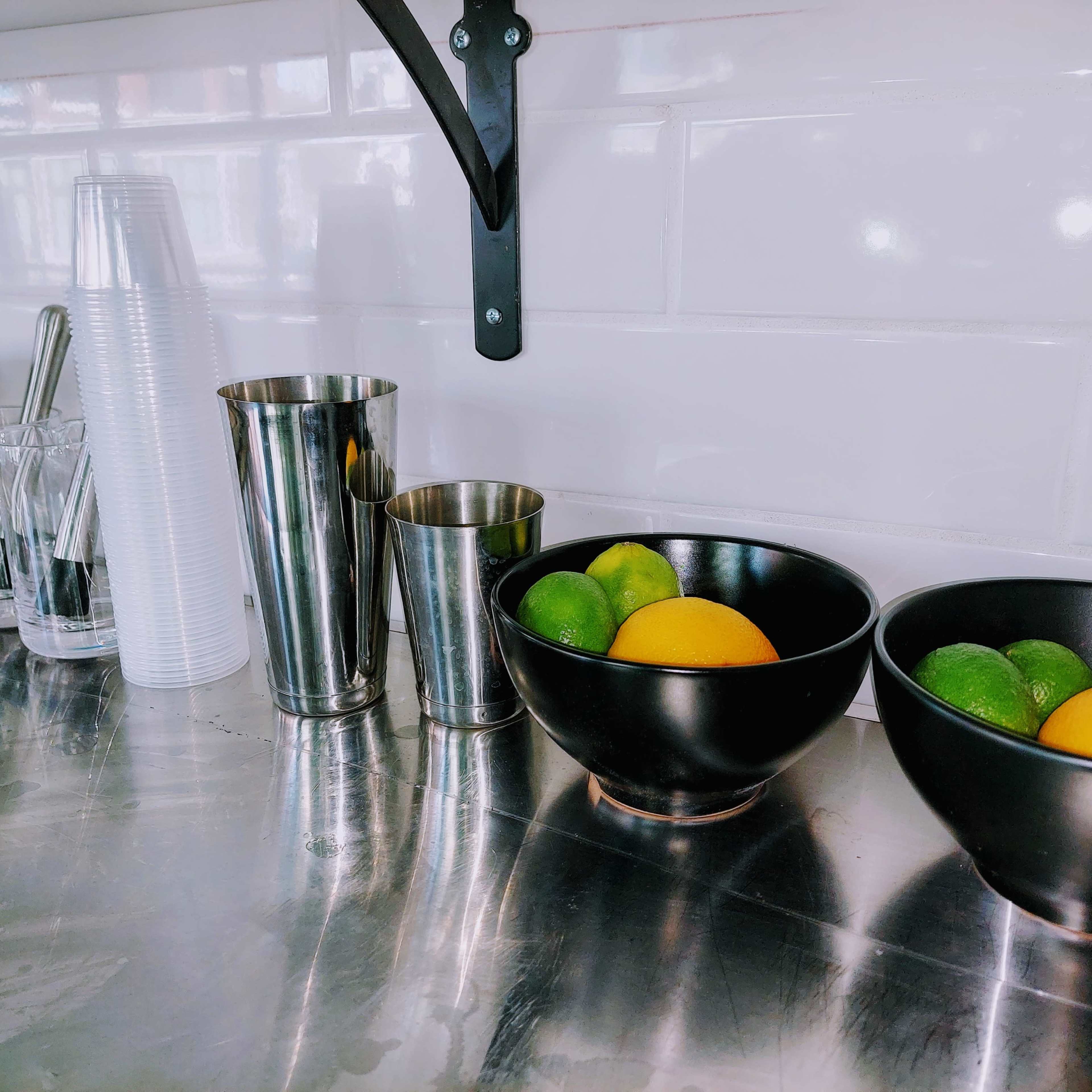 A countertop with various metallic cups and glasses alongside two bowls containing oranges and limes.