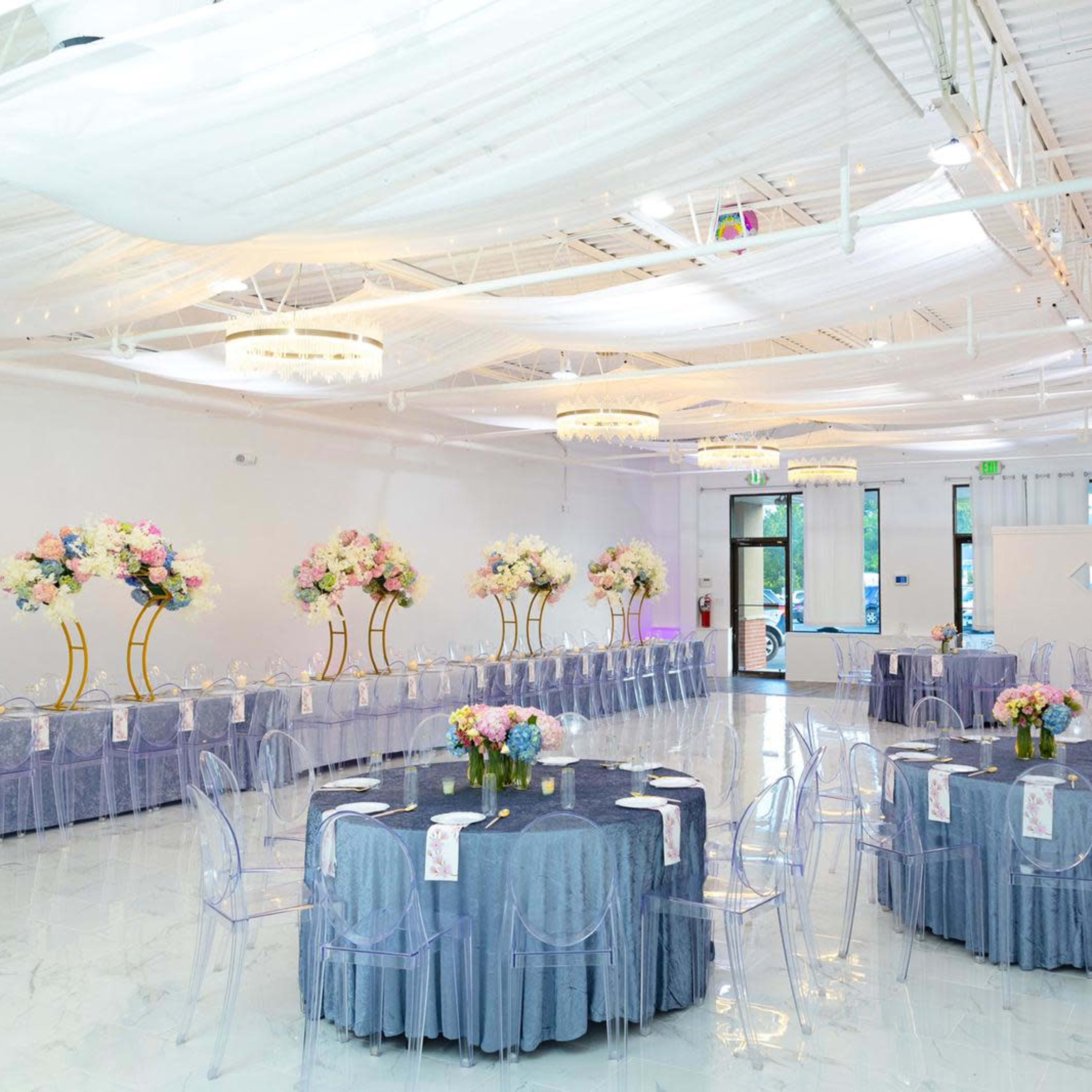 A decorated event space with round tables covered in lavender tablecloths and floral arrangements, surrounded by transparent chairs and soft lighting.