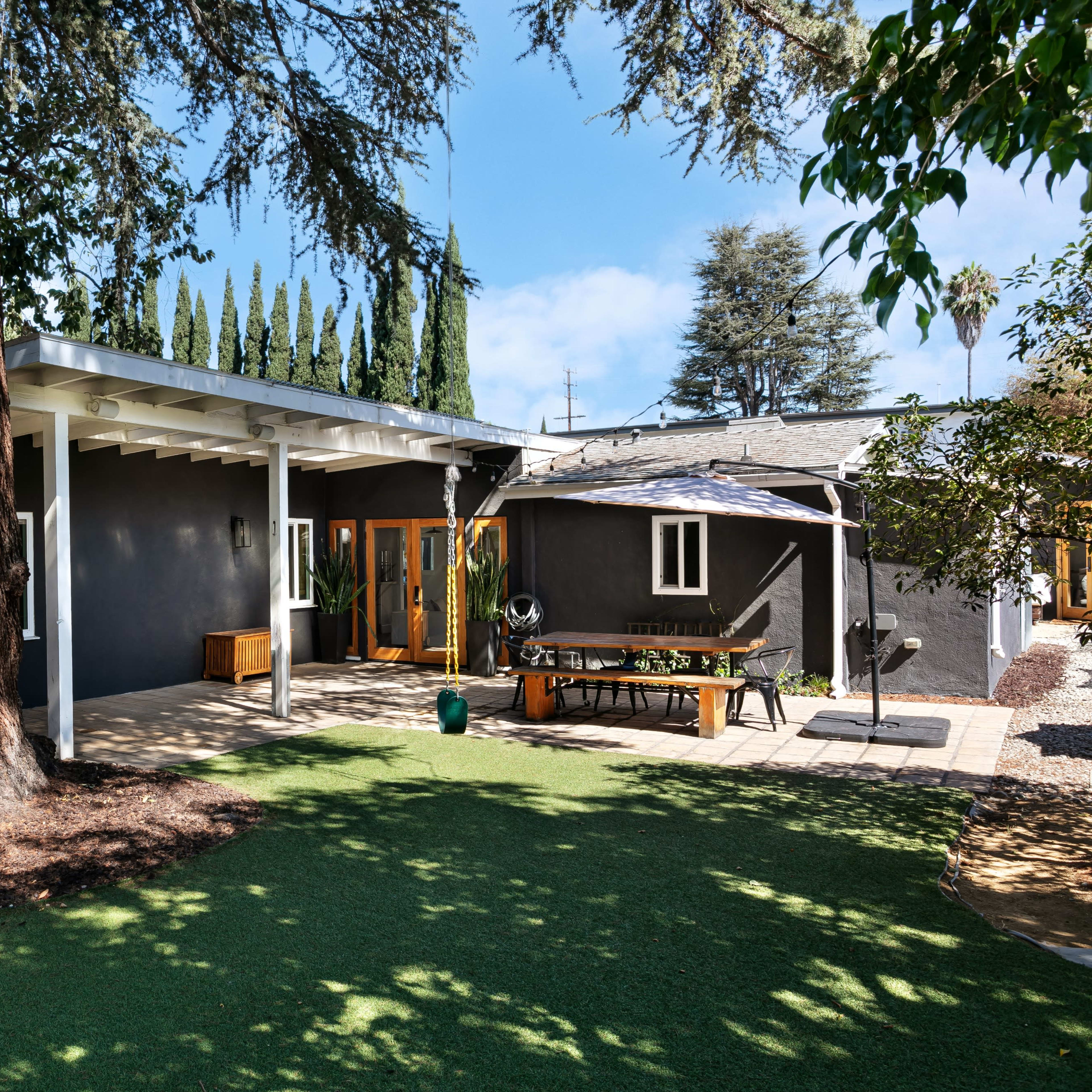 A modern single-story house with a black exterior, surrounded by greenery and a patio area featuring a table and benches.