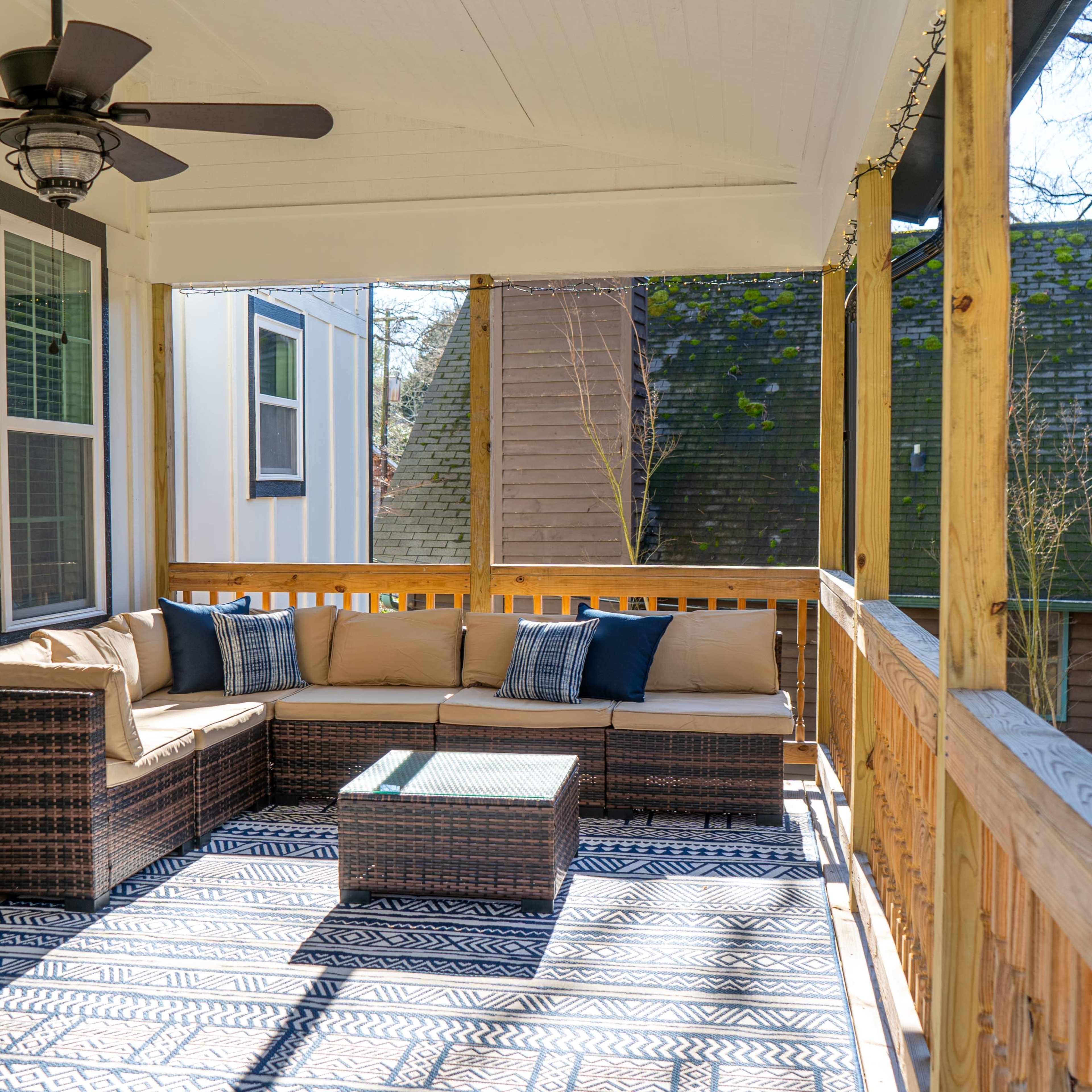 A covered porch features a wicker seating area with cushions and a coffee table, surrounded by wooden railings and large windows.
