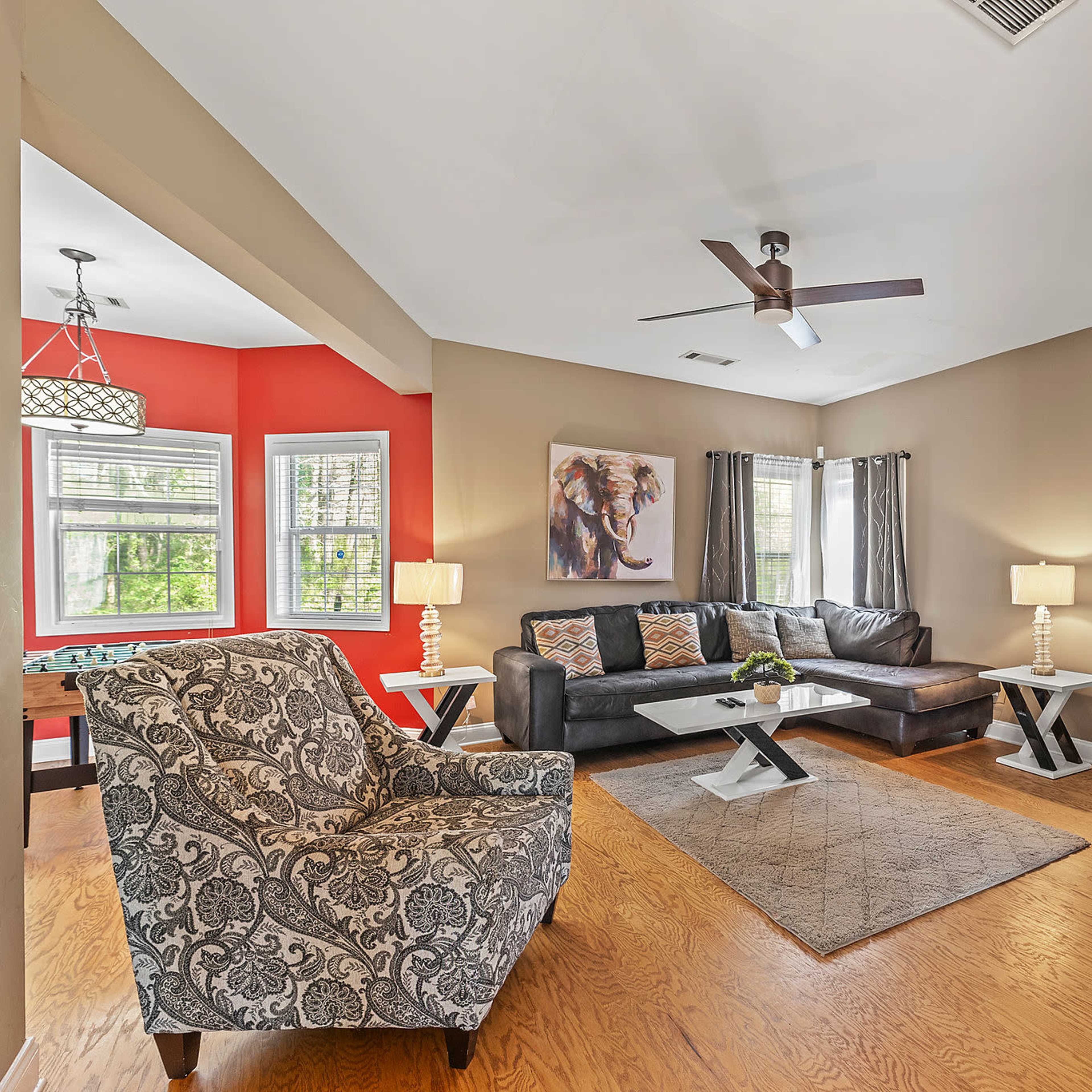 The living room features a black leather sofa, a patterned chair, and a coffee table, with walls painted in beige and red, and large windows allowing natural light.