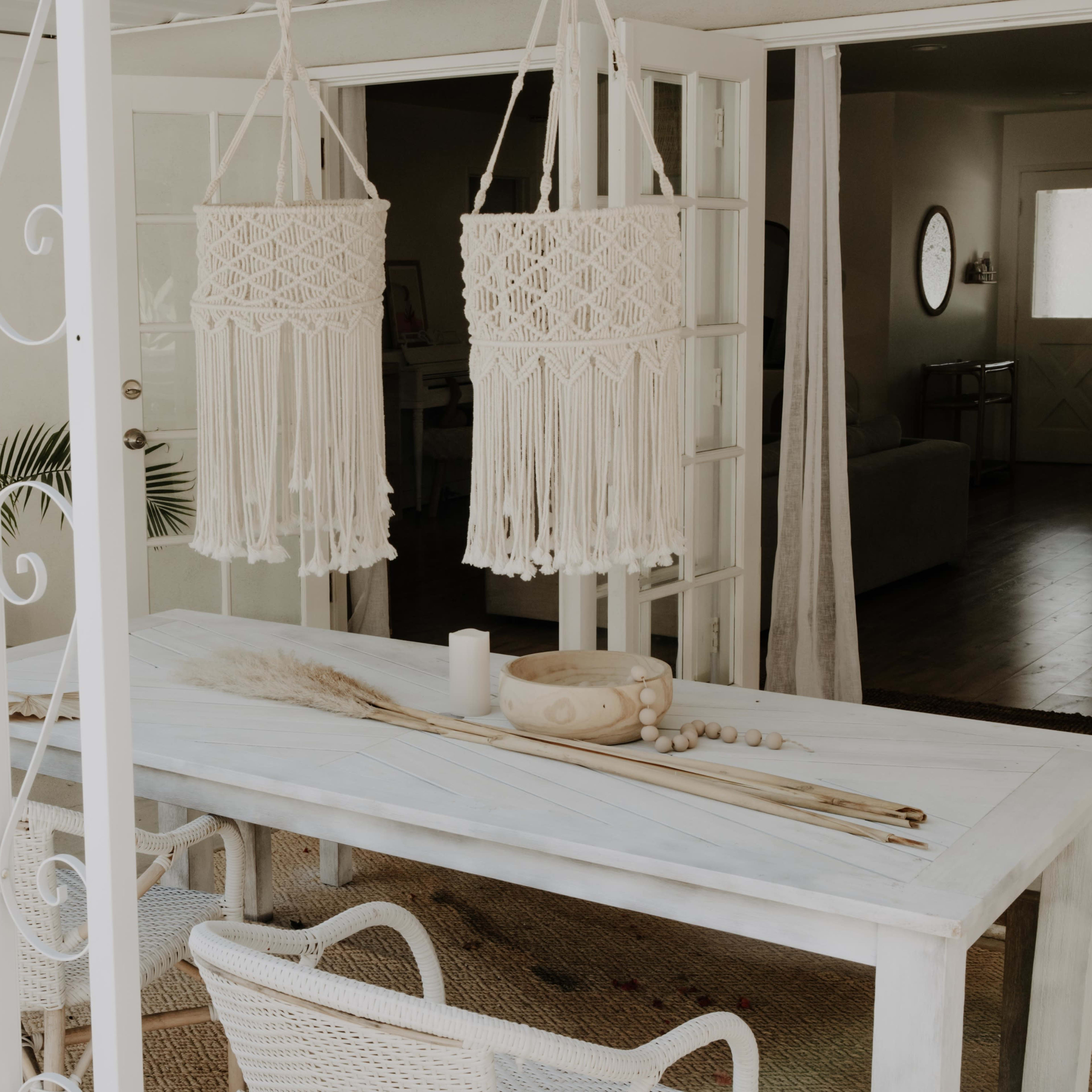 A white table is set outdoors with decorative items, while two macramé planters hang above it and a mirror leans against the wall.