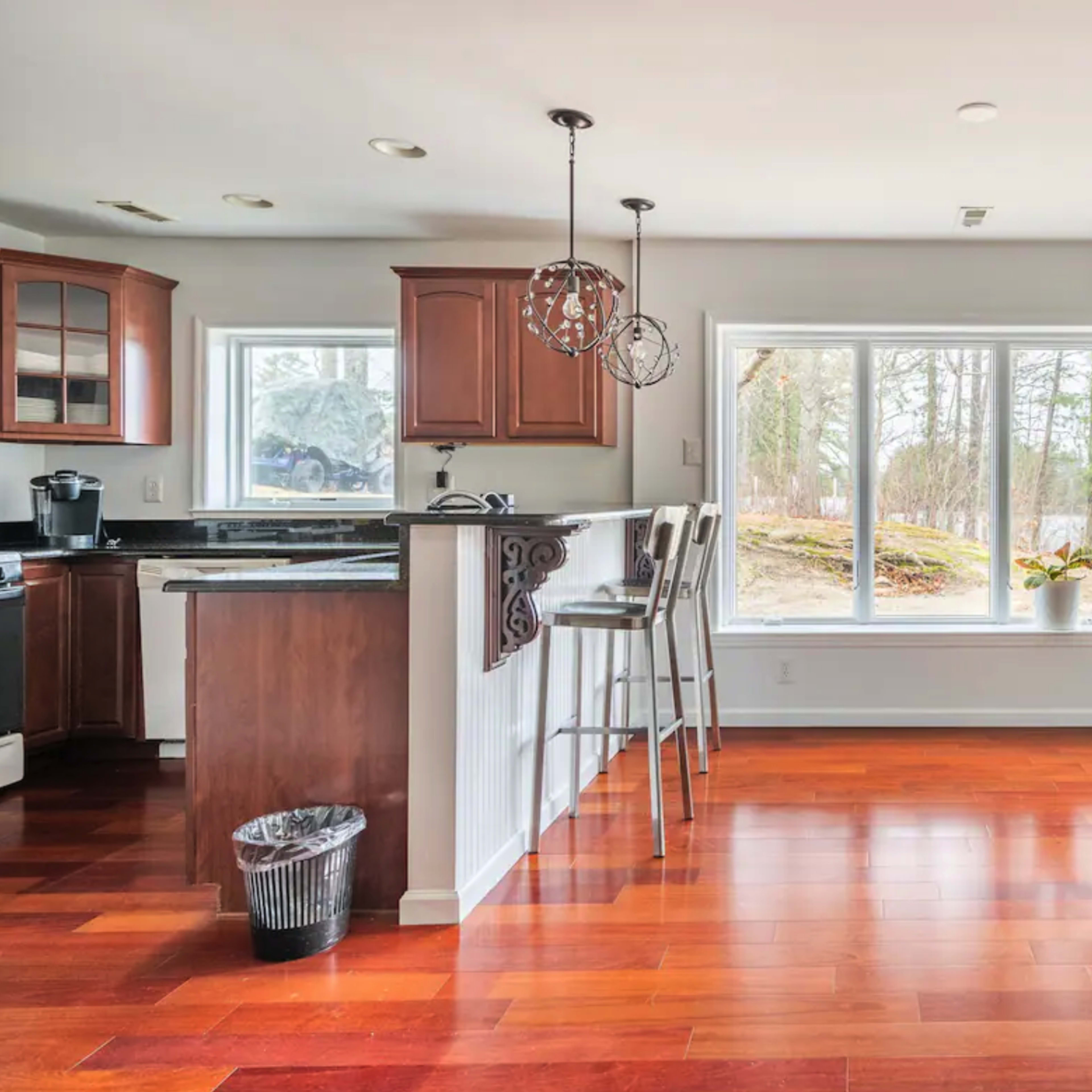 A modern kitchen features wooden cabinets, a bar with stools, and large windows overlooking a wooded area.