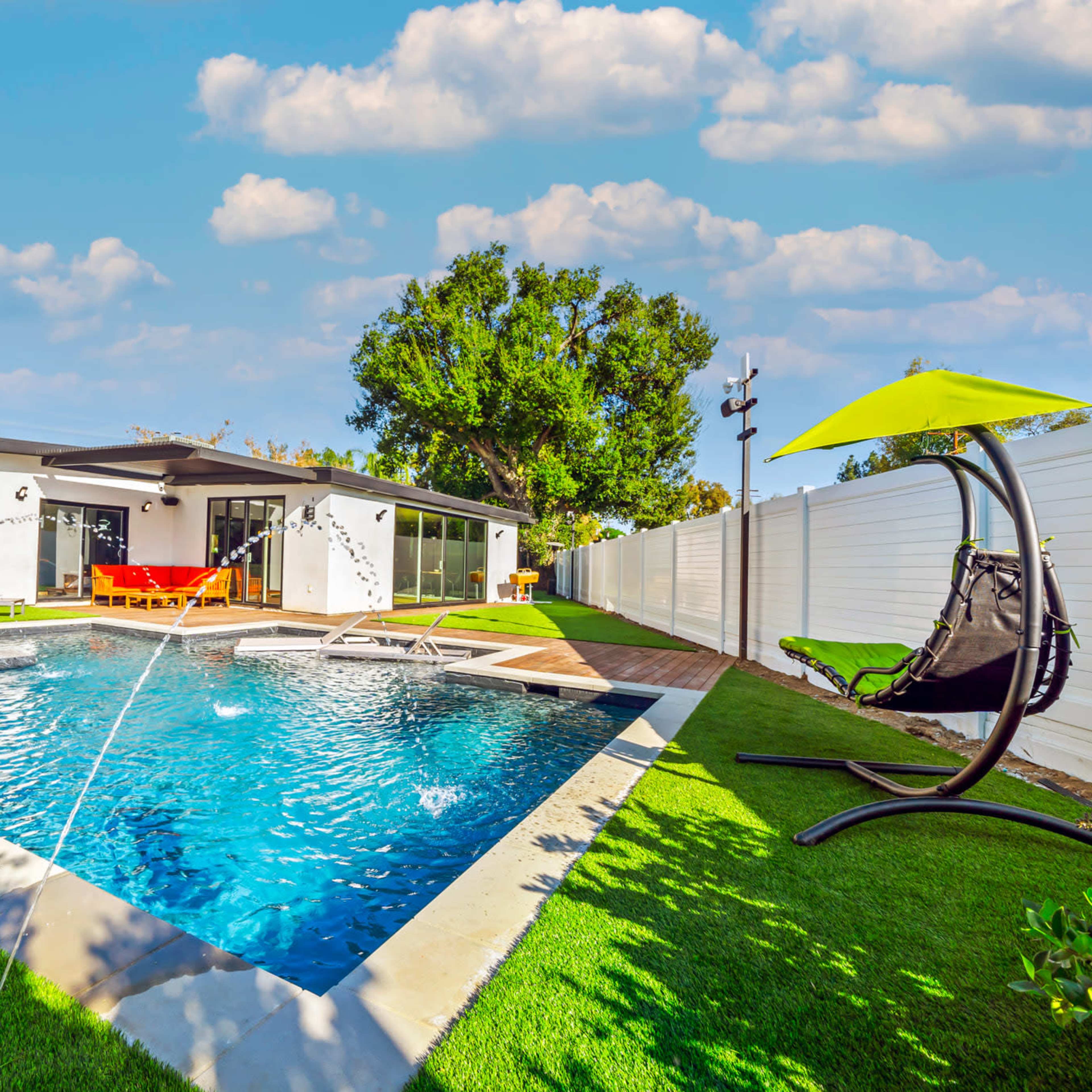 A modern backyard featuring a swimming pool with water fountains, a hanging chair, and a house in the background, surrounded by green grass and white fencing.