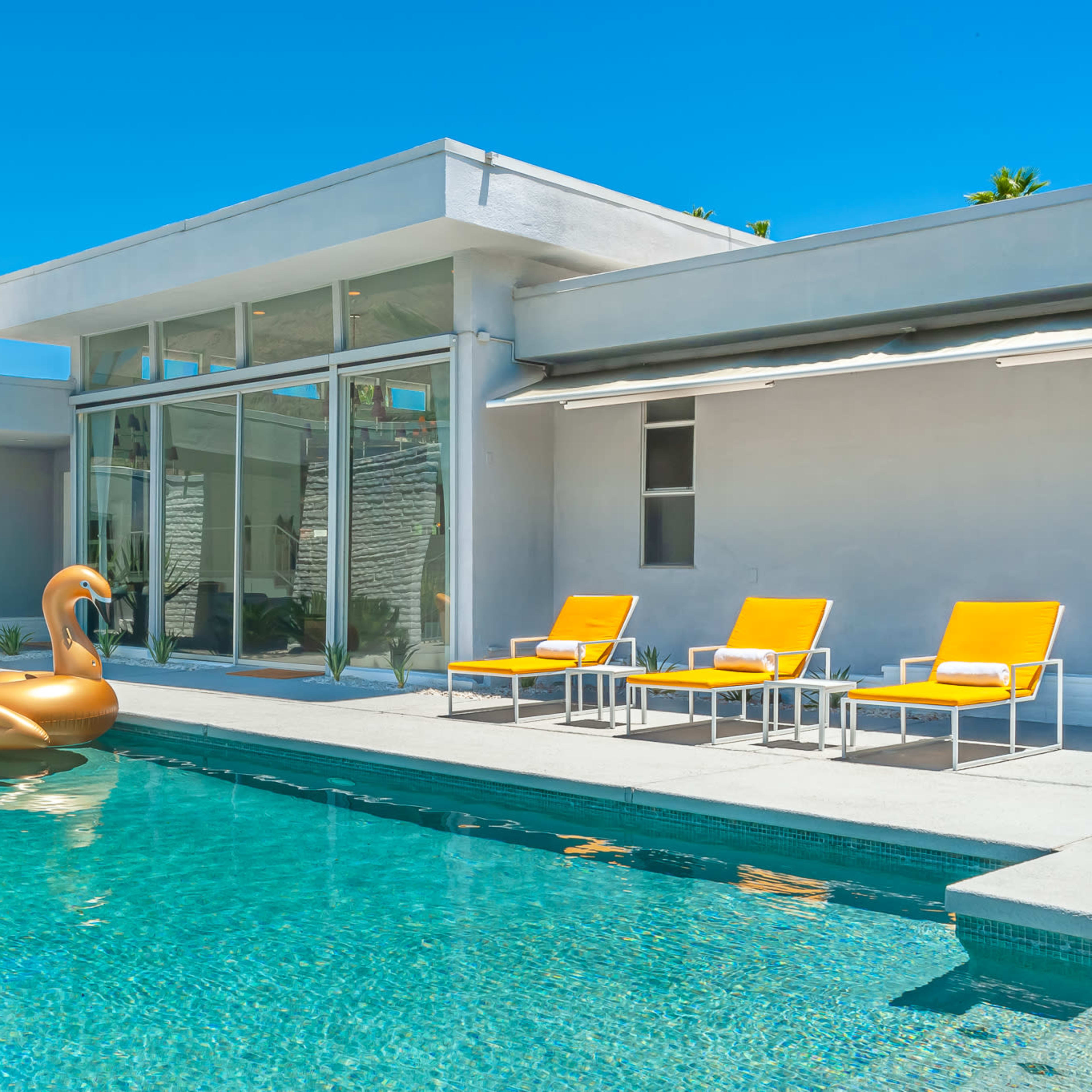 A modern pool area features bright orange lounge chairs, a large inflatable swan, and glass walls that allow for views into the interior spaces.
