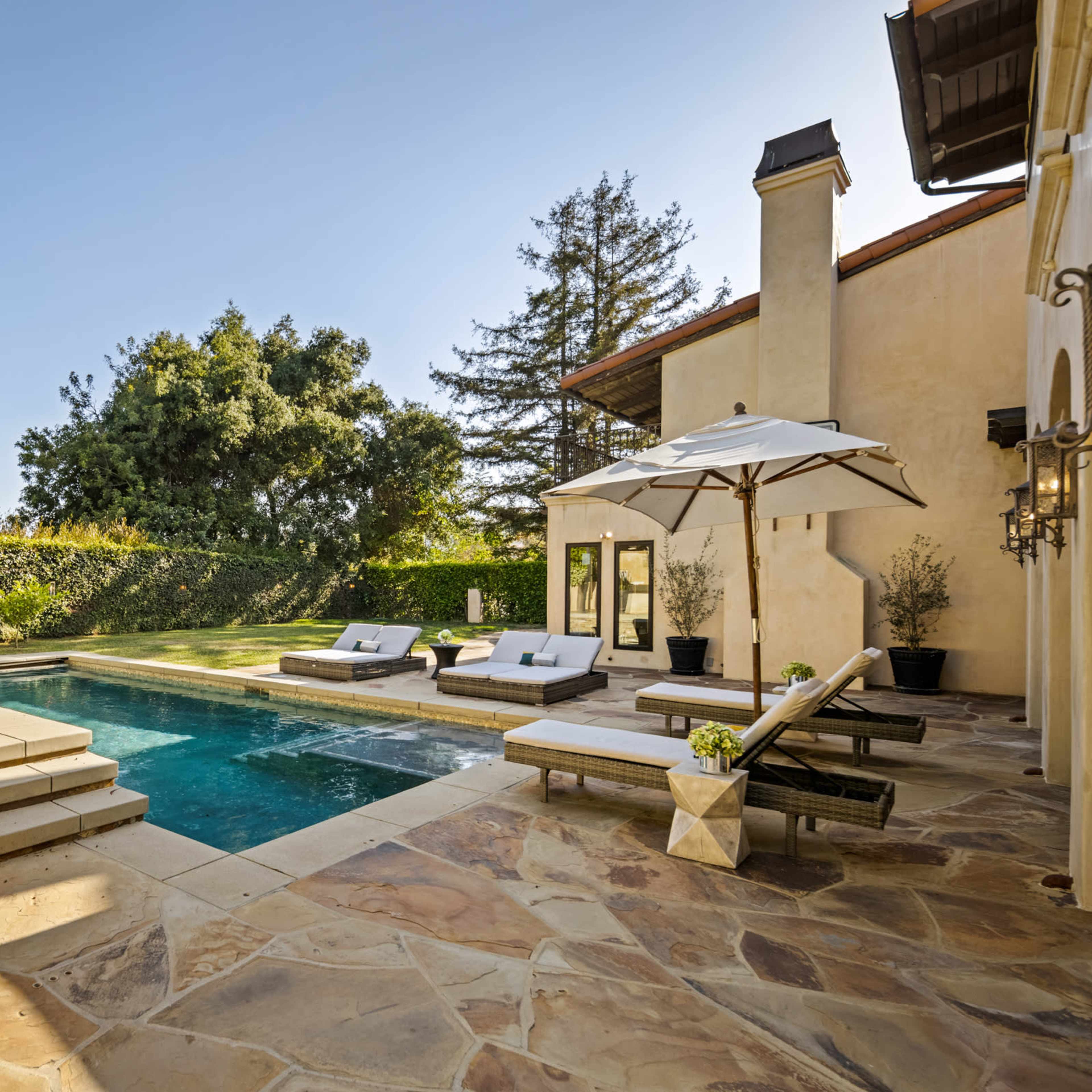 A residential backyard features a pool with lounge chairs, surrounded by landscaped greenery and a patio area with an umbrella.