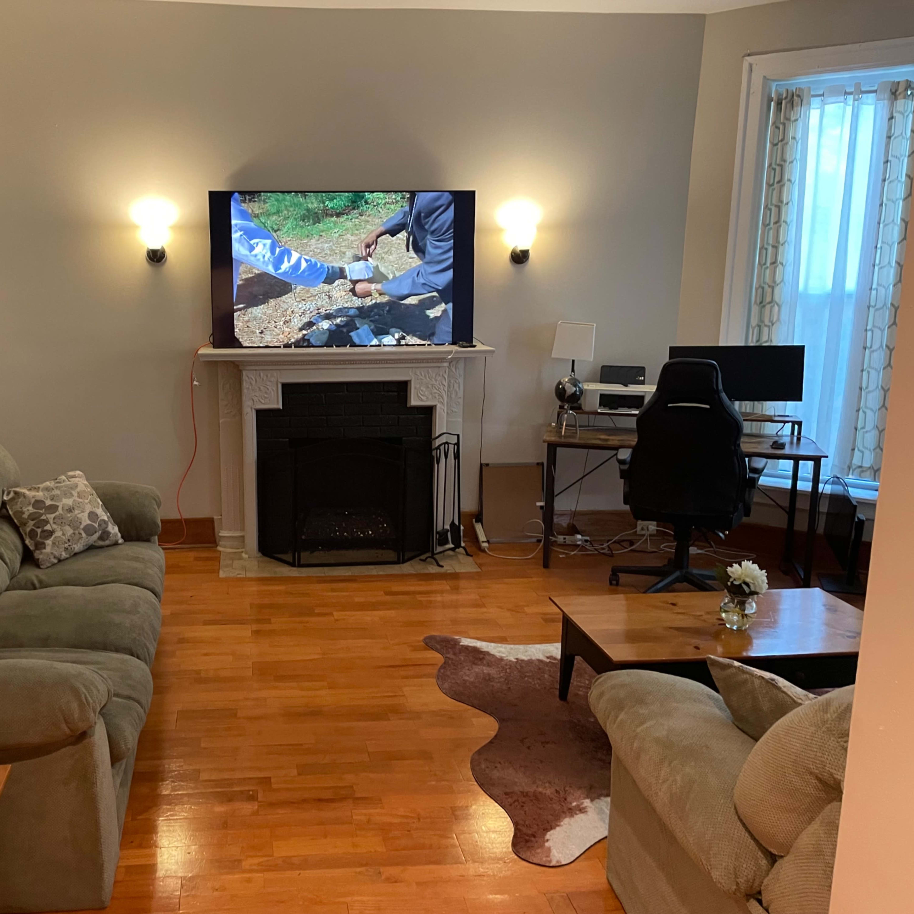 A living room features a couch, armchair, wall-mounted TV above a fireplace, and a desk with a chair.