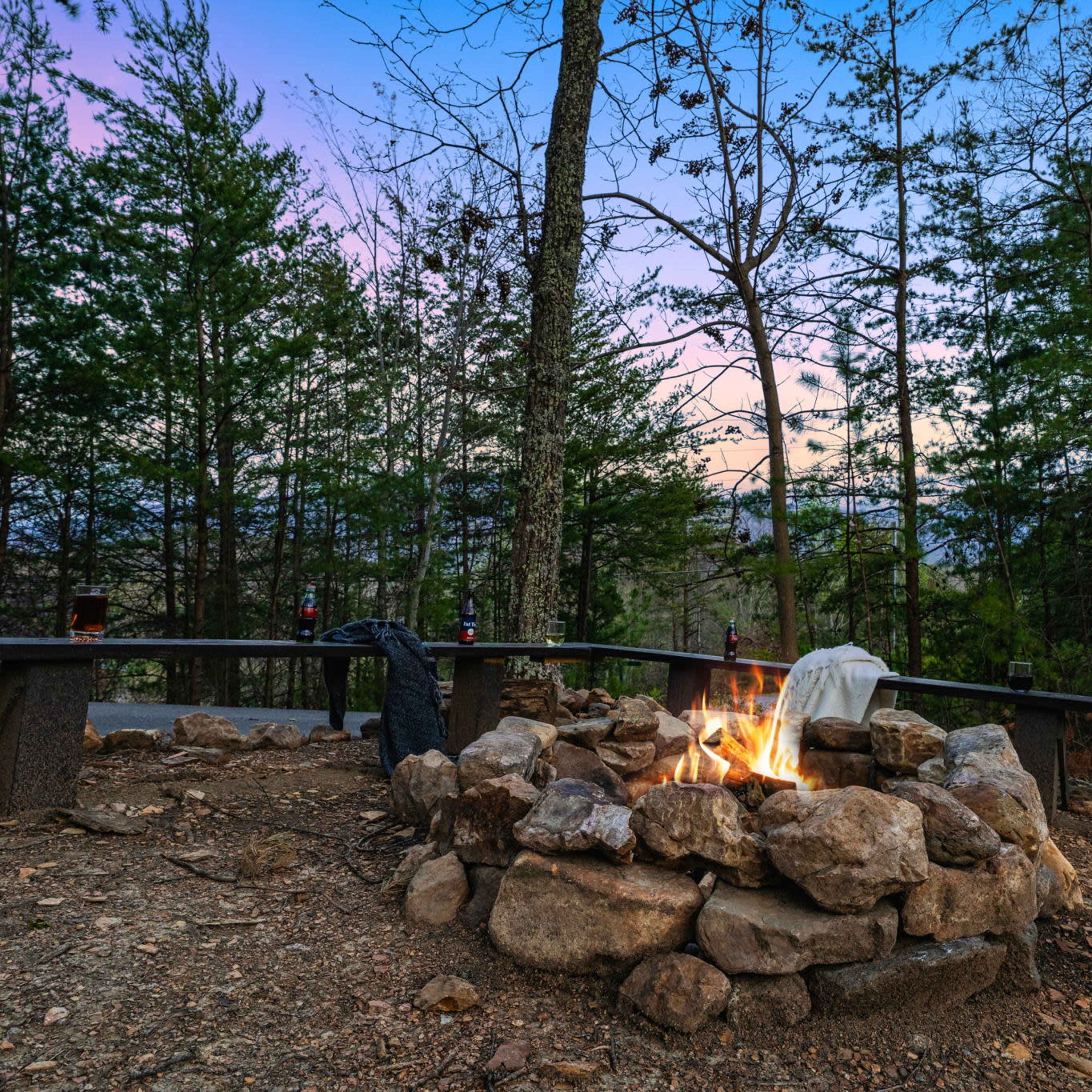 A campfire sits within a stone circle surrounded by trees at dusk, with a wooden bench nearby.