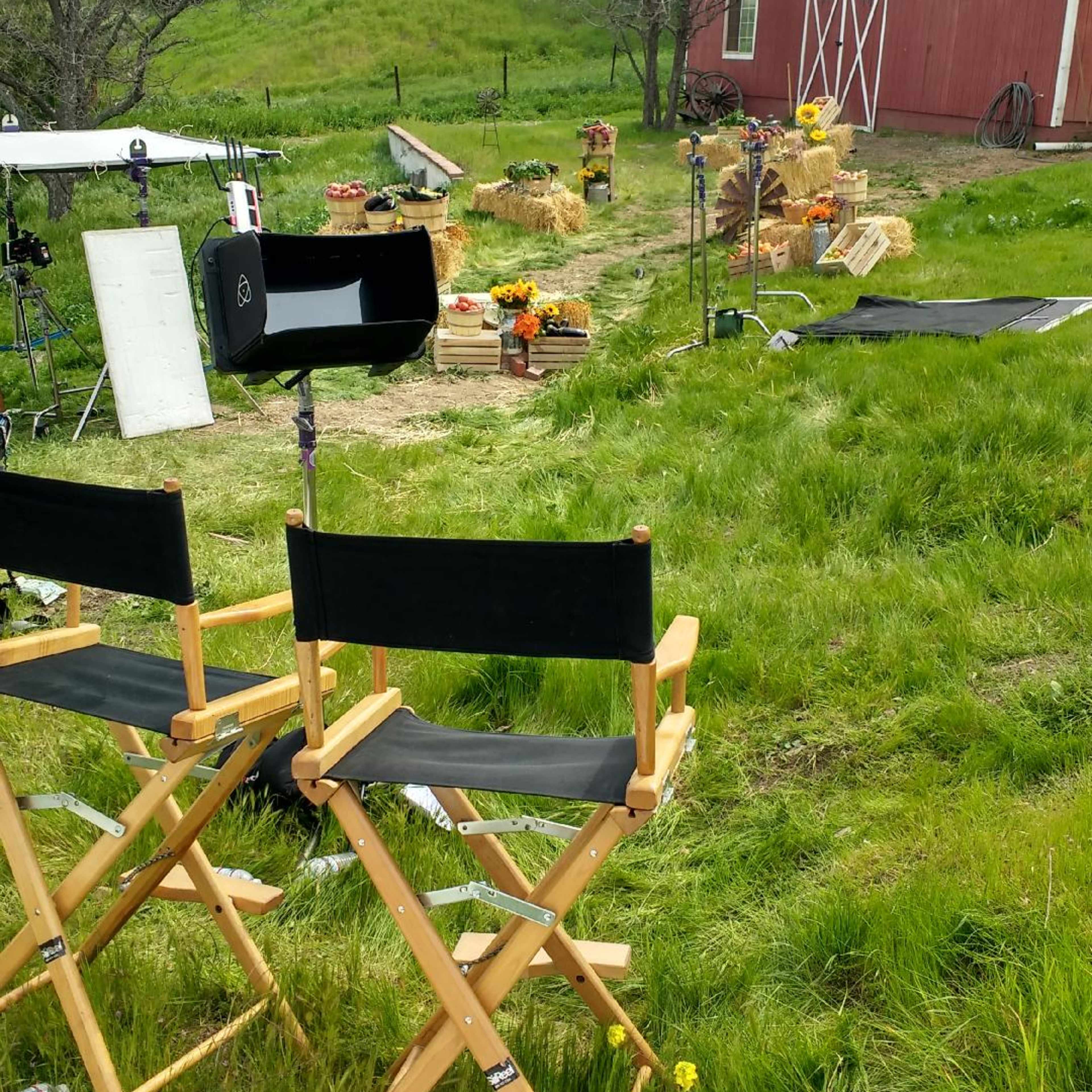 A film set in a grassy area with director's chairs facing a scene decorated with hay bales and flowers near a red barn.