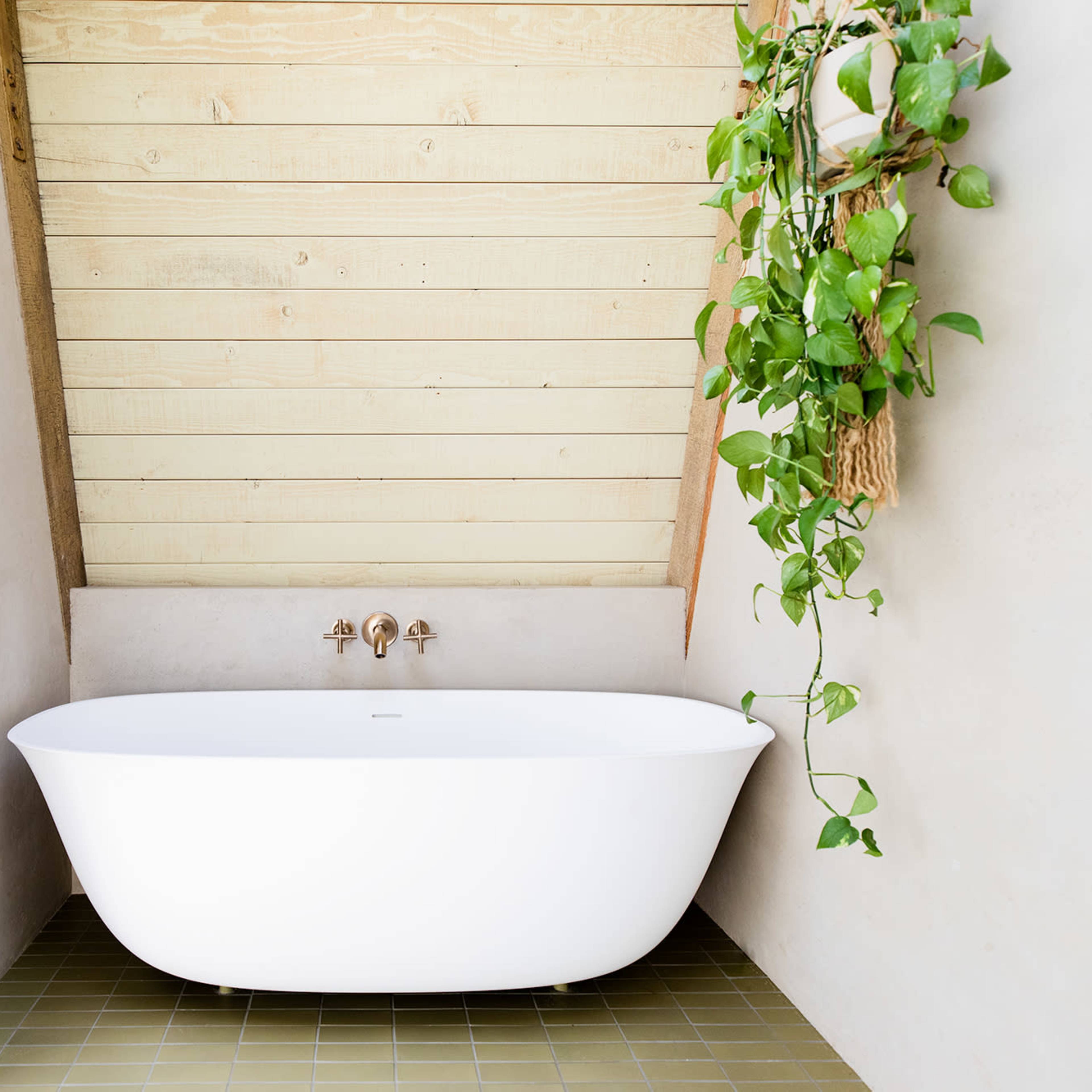 A white freestanding bathtub is positioned in a corner of a room with slanted wooden walls and a small green plant hanging nearby.