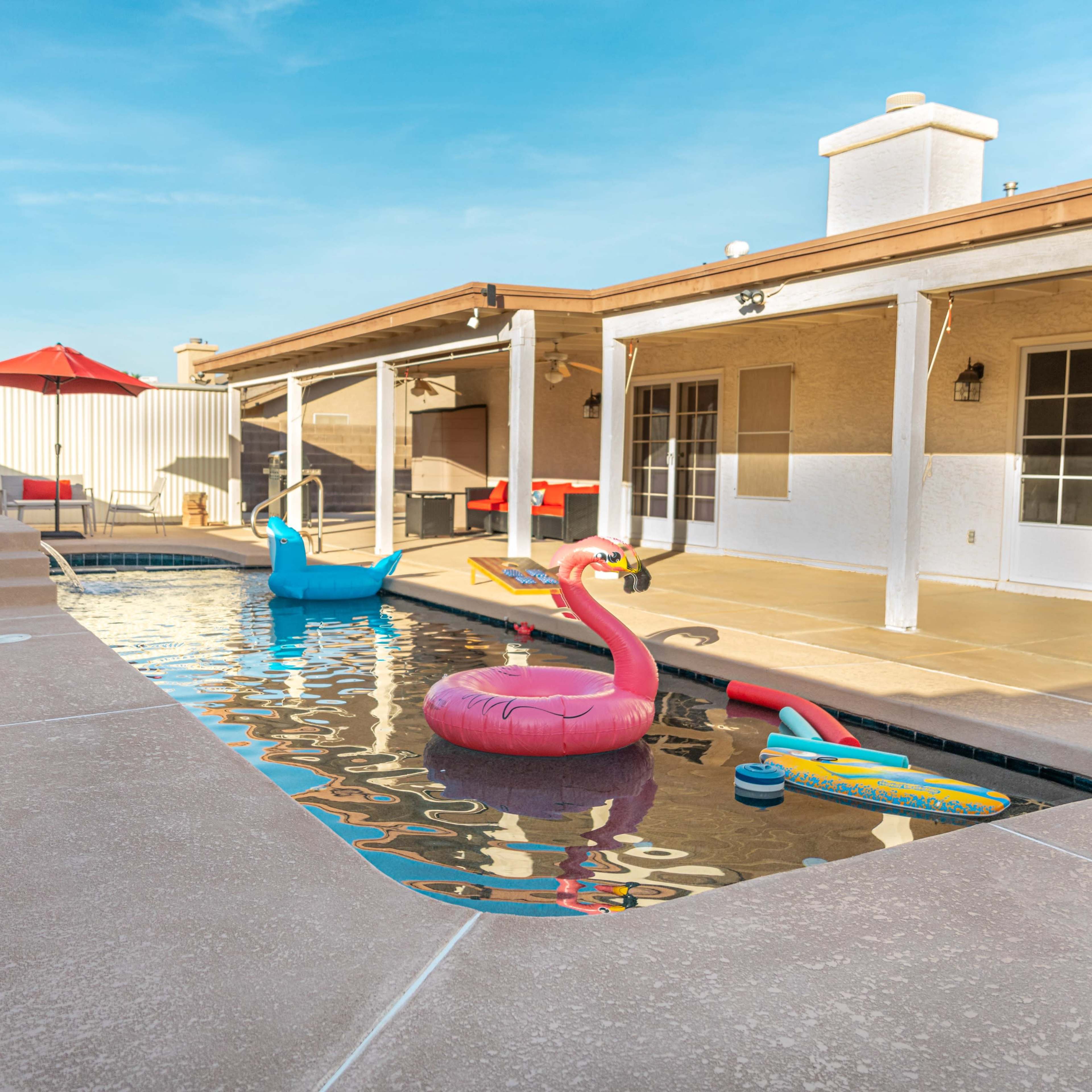 A swimming pool with colorful floats, surrounded by a patio and a house, under a clear blue sky.