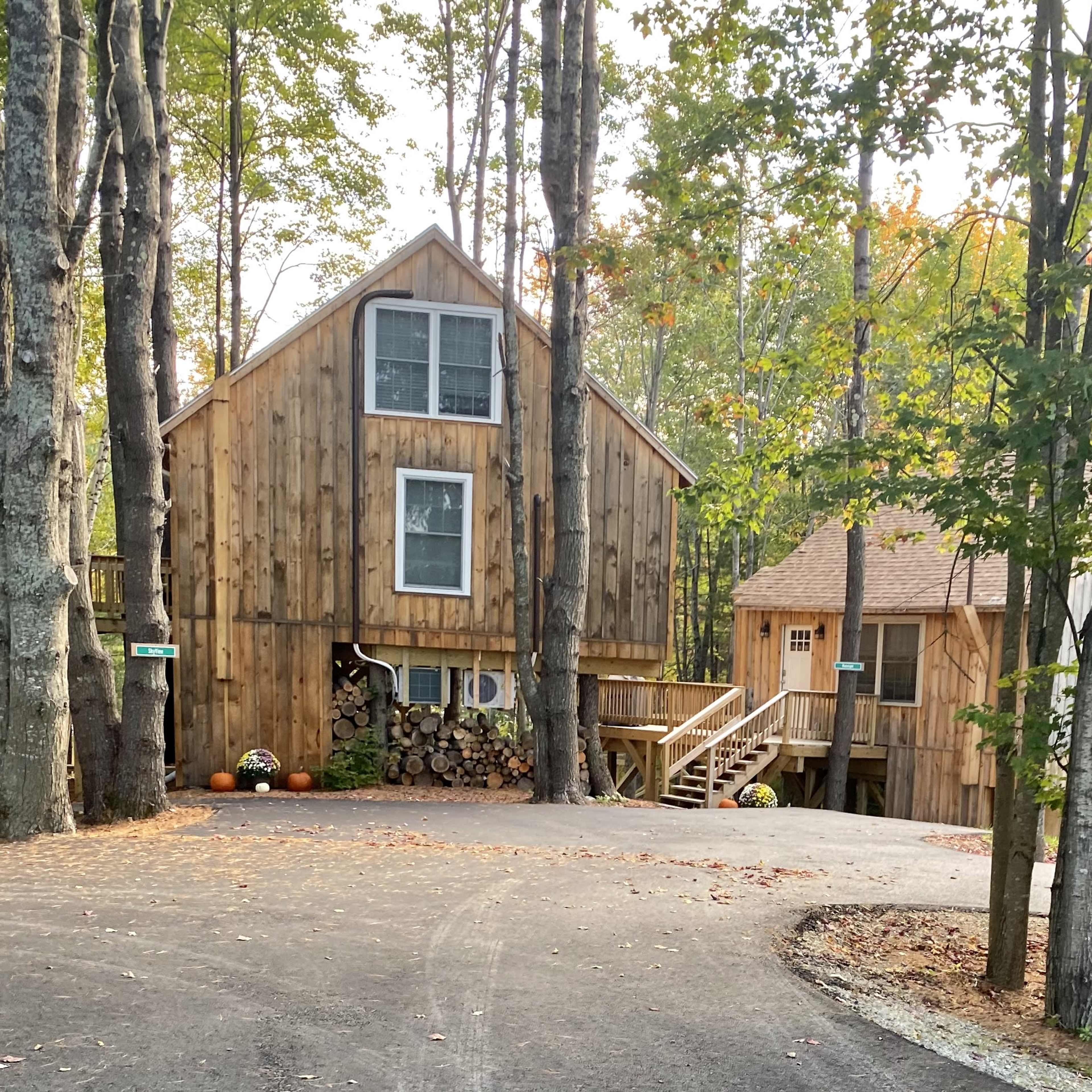 A wooden house with large windows sits among trees, featuring a driveway that curves around to the entrance.