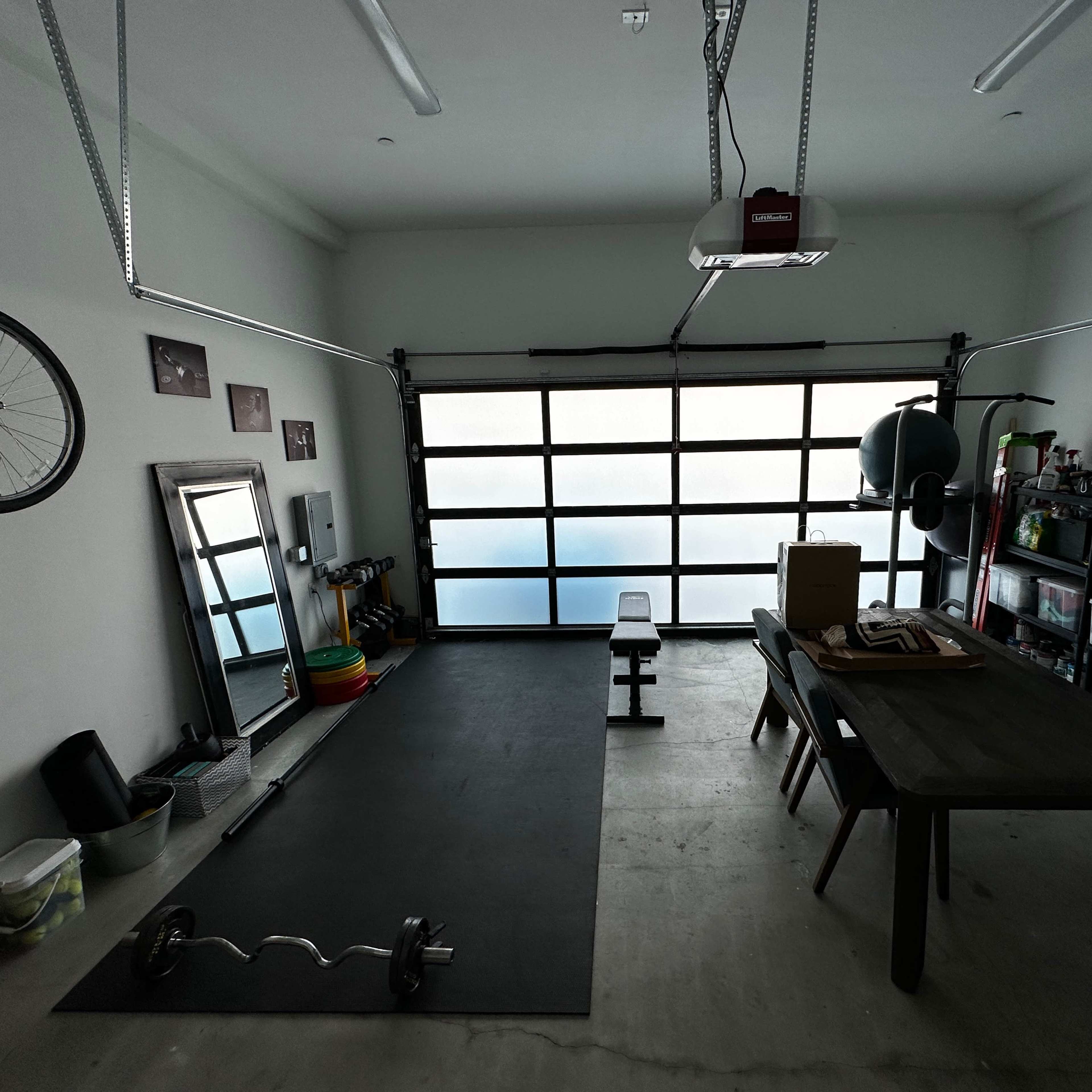 A garage space features workout equipment, a table, storage shelves, a mirror, and a bicycle hung on the wall.