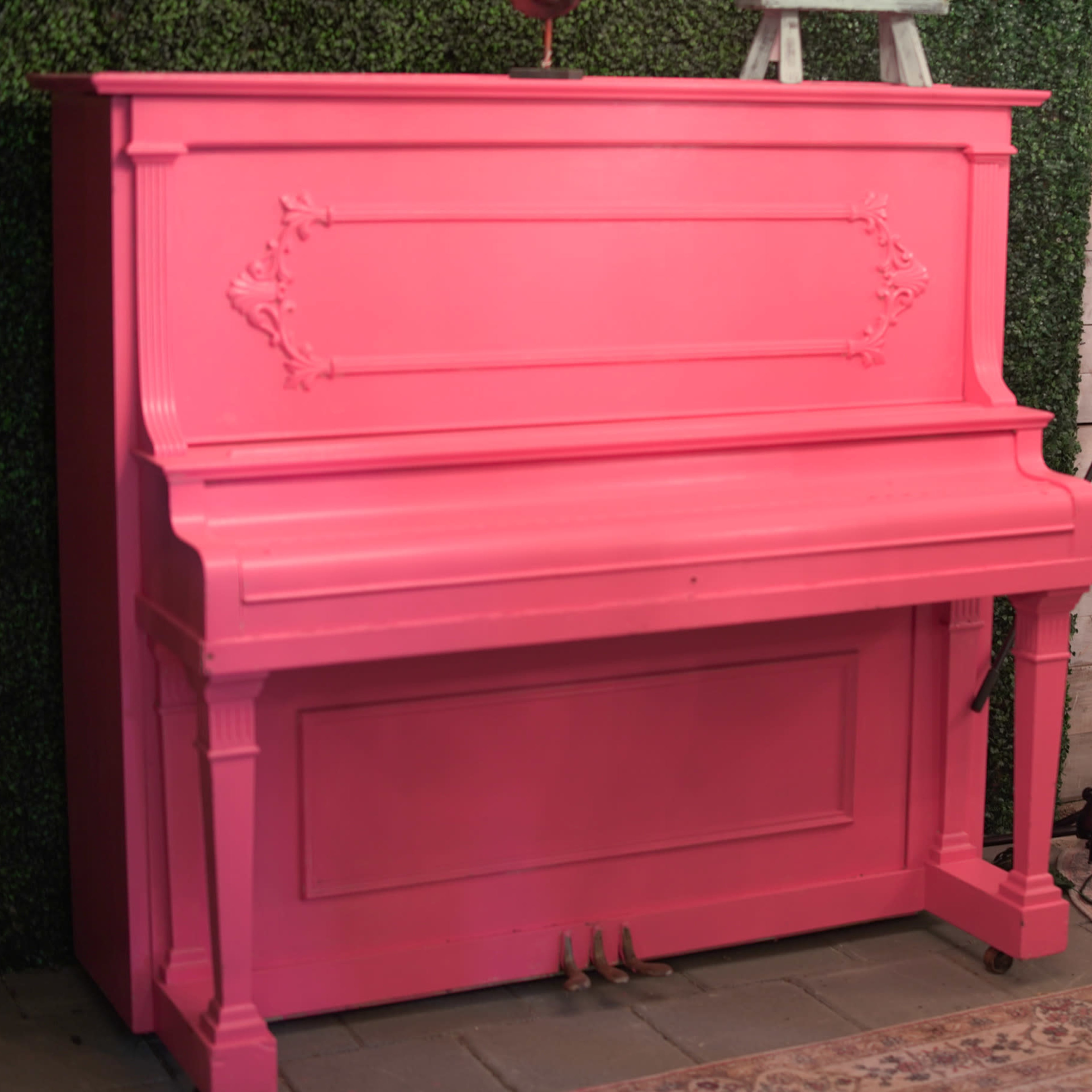 A pink upright piano stands against a backdrop of green foliage.