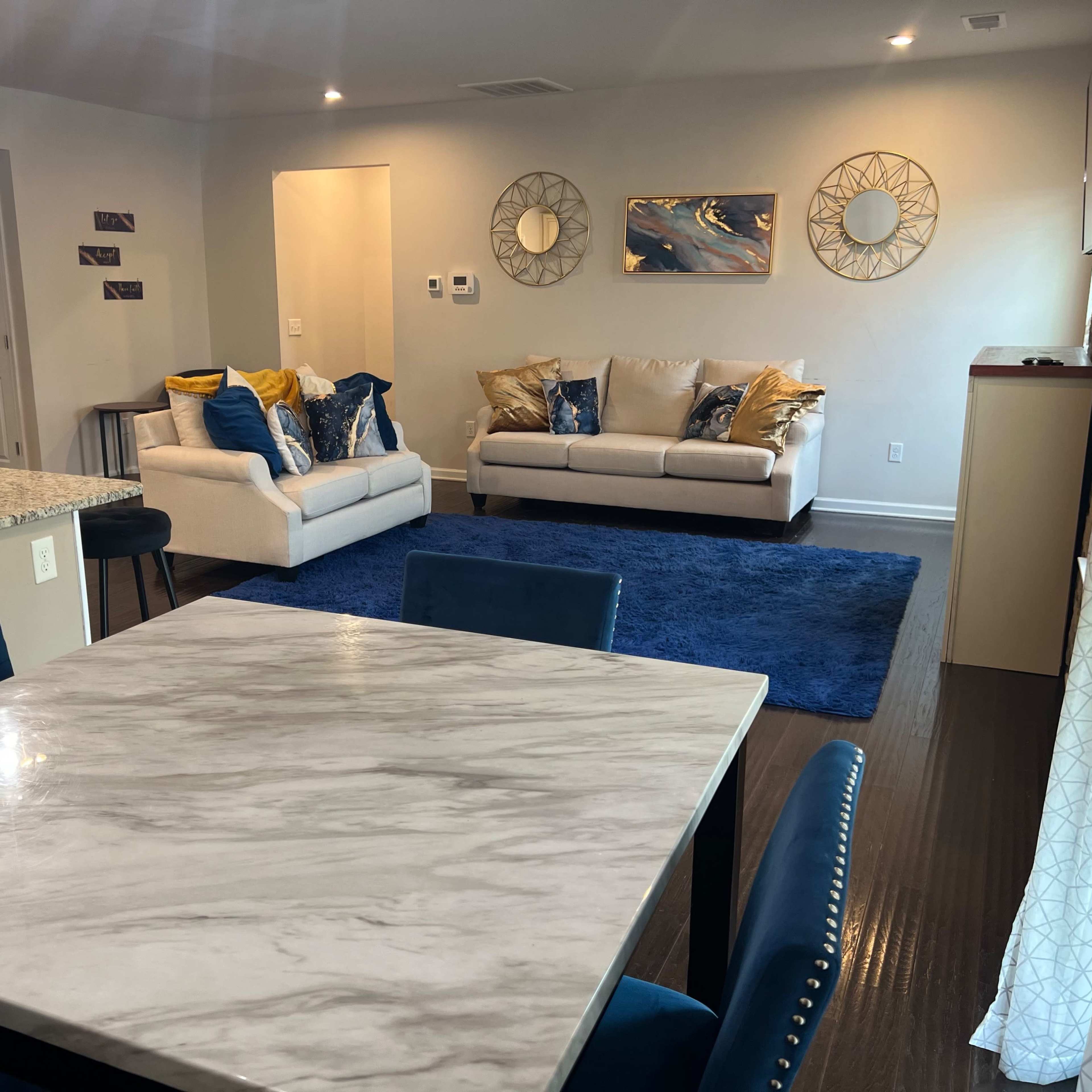 A modern living room and dining area featuring a marble dining table, blue upholstered chairs, two sofas with decorative pillows, and a blue rug on a wooden floor.