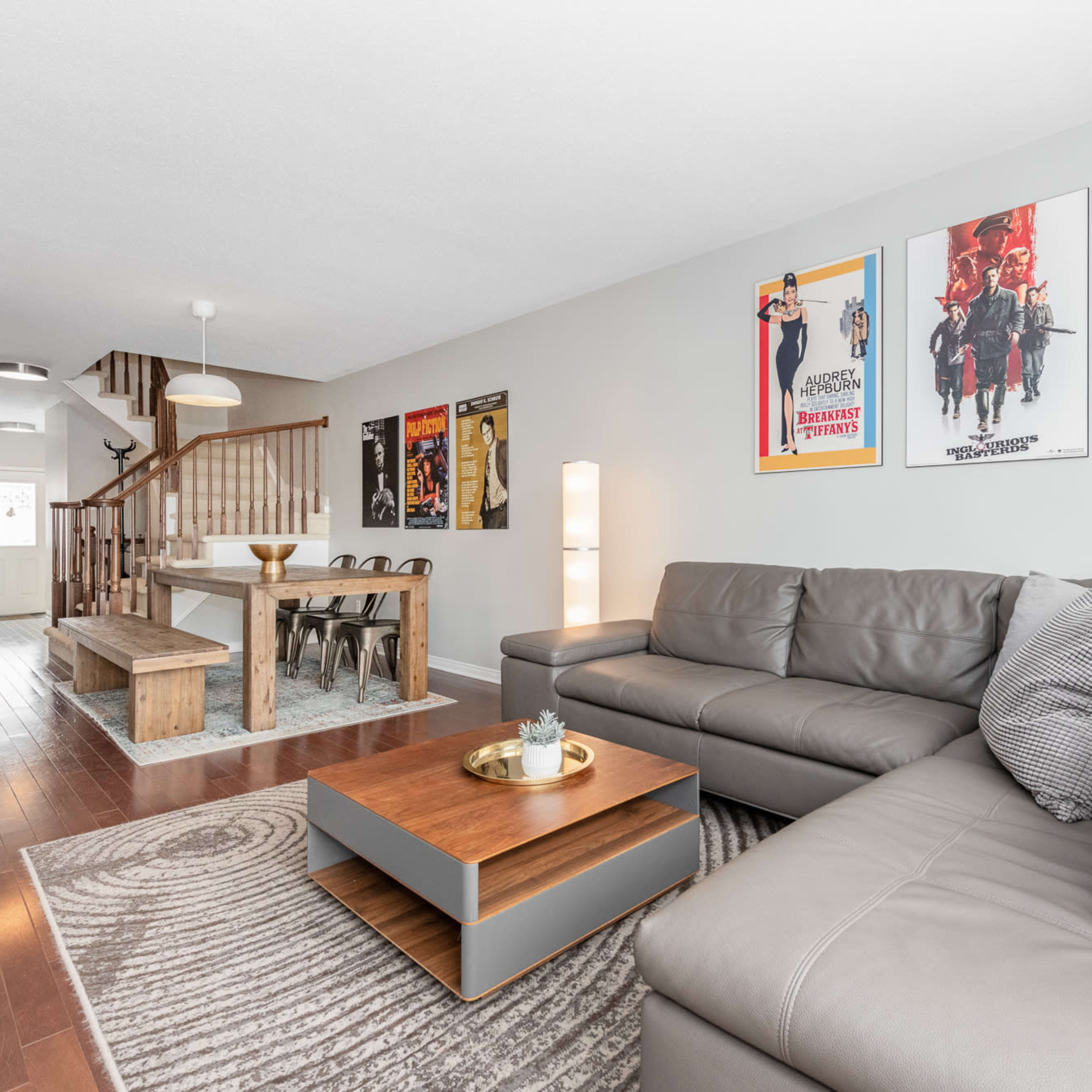 A modern living room with a gray sectional sofa, a wooden coffee table, and vintage movie posters on the walls, adjacent to a staircase leading to an upper level.