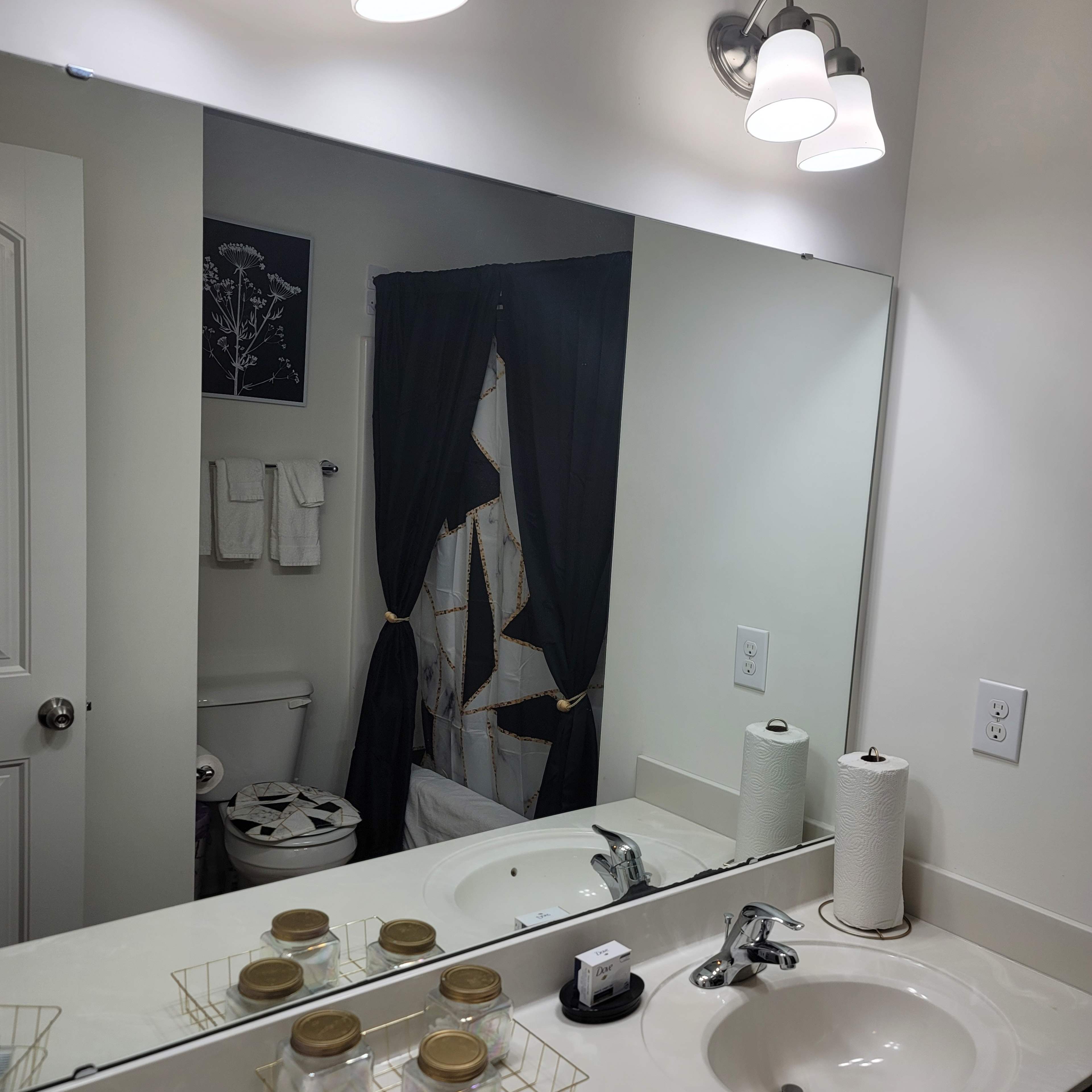 A bathroom sink area with a mirror, light fixtures above, and a decorative curtain partially obscuring the view of the shower and toilet.