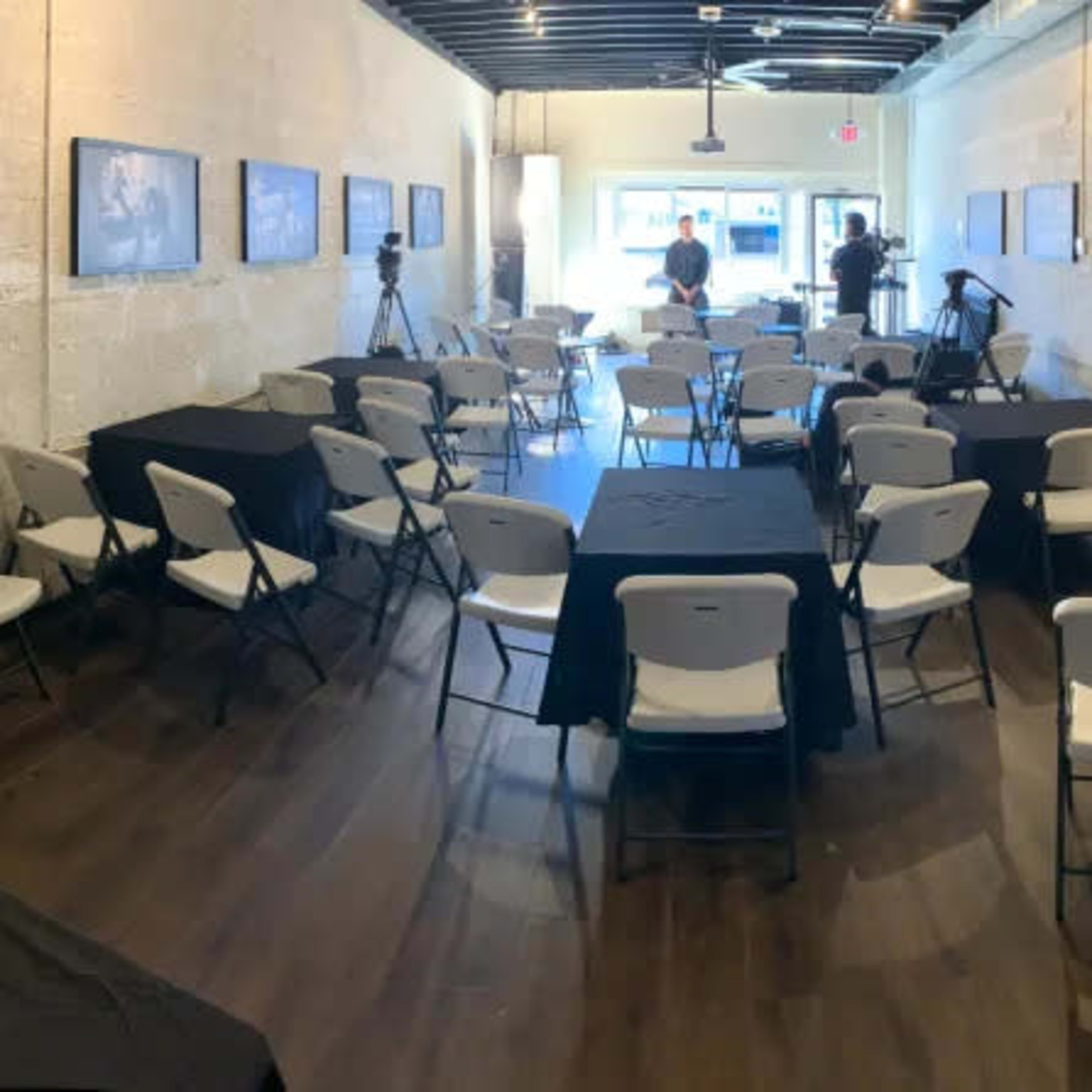 A spacious interior room arranged with rows of white chairs and black tables, with black-and-white photographs hanging on the walls.