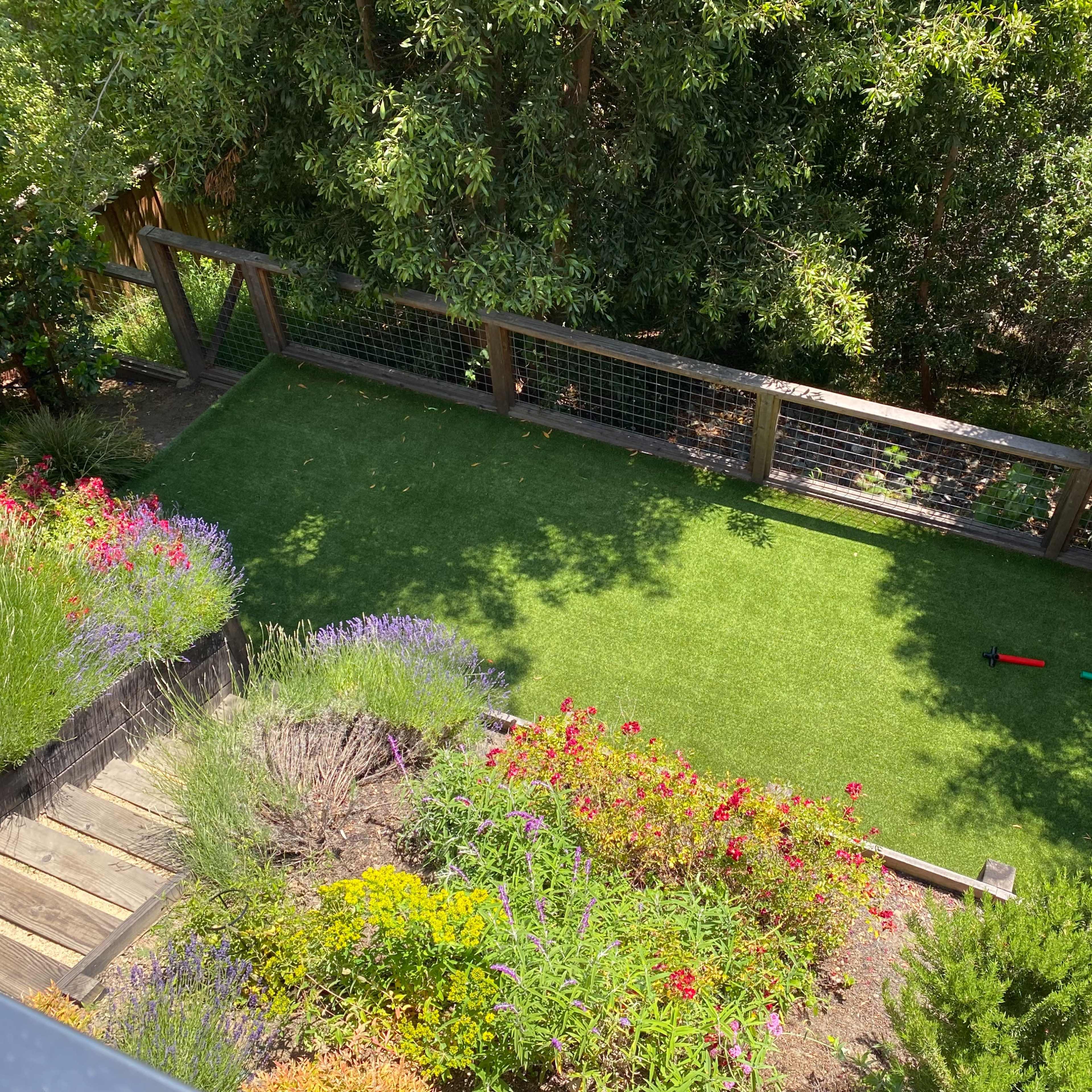 An aerial view of a landscaped backyard with artificial grass surrounded by colorful flower beds and a staircase leading down to the area.