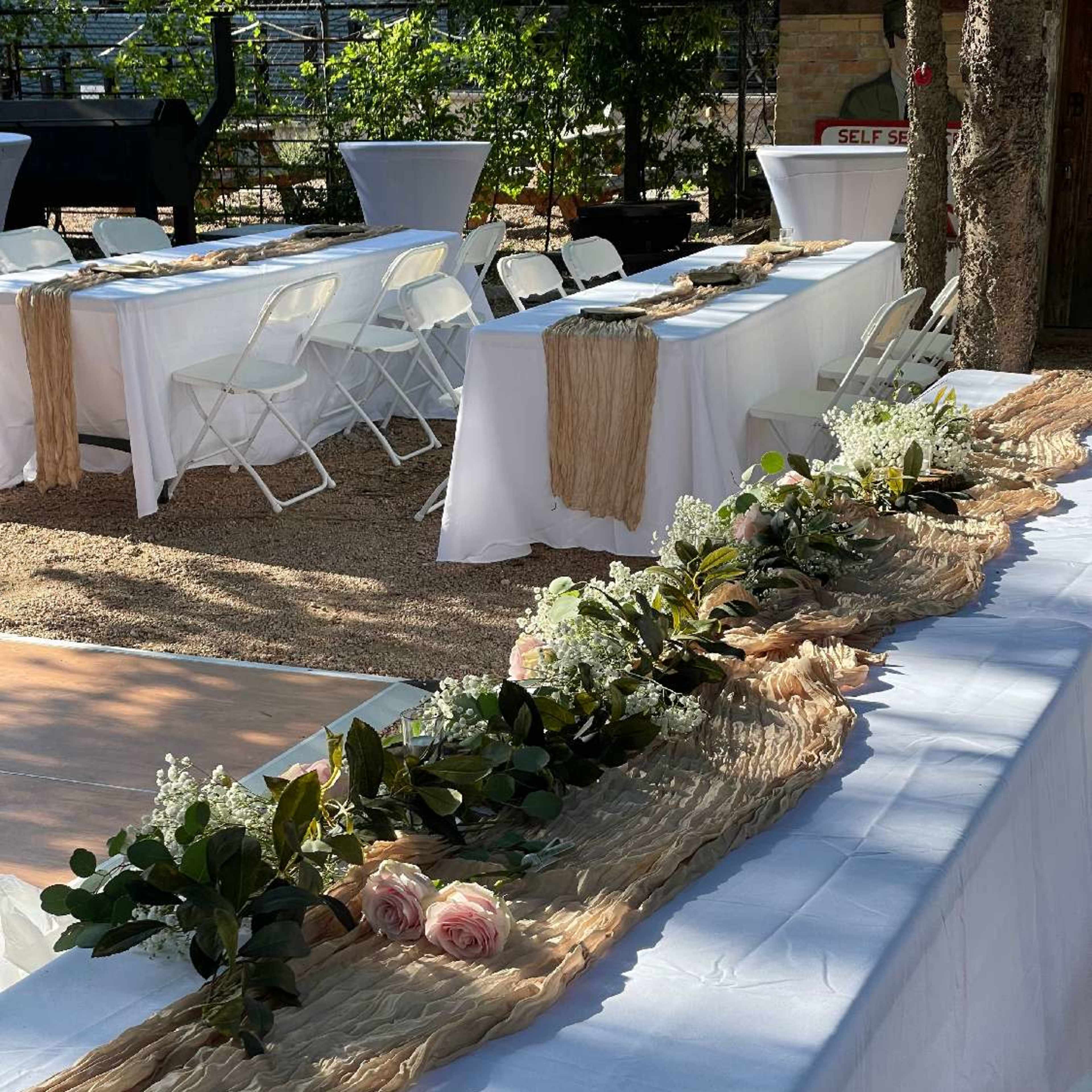 A set-up for an outdoor event with several tables covered in white linens, adorned with a floral centerpiece of roses and greenery along the length of the table.