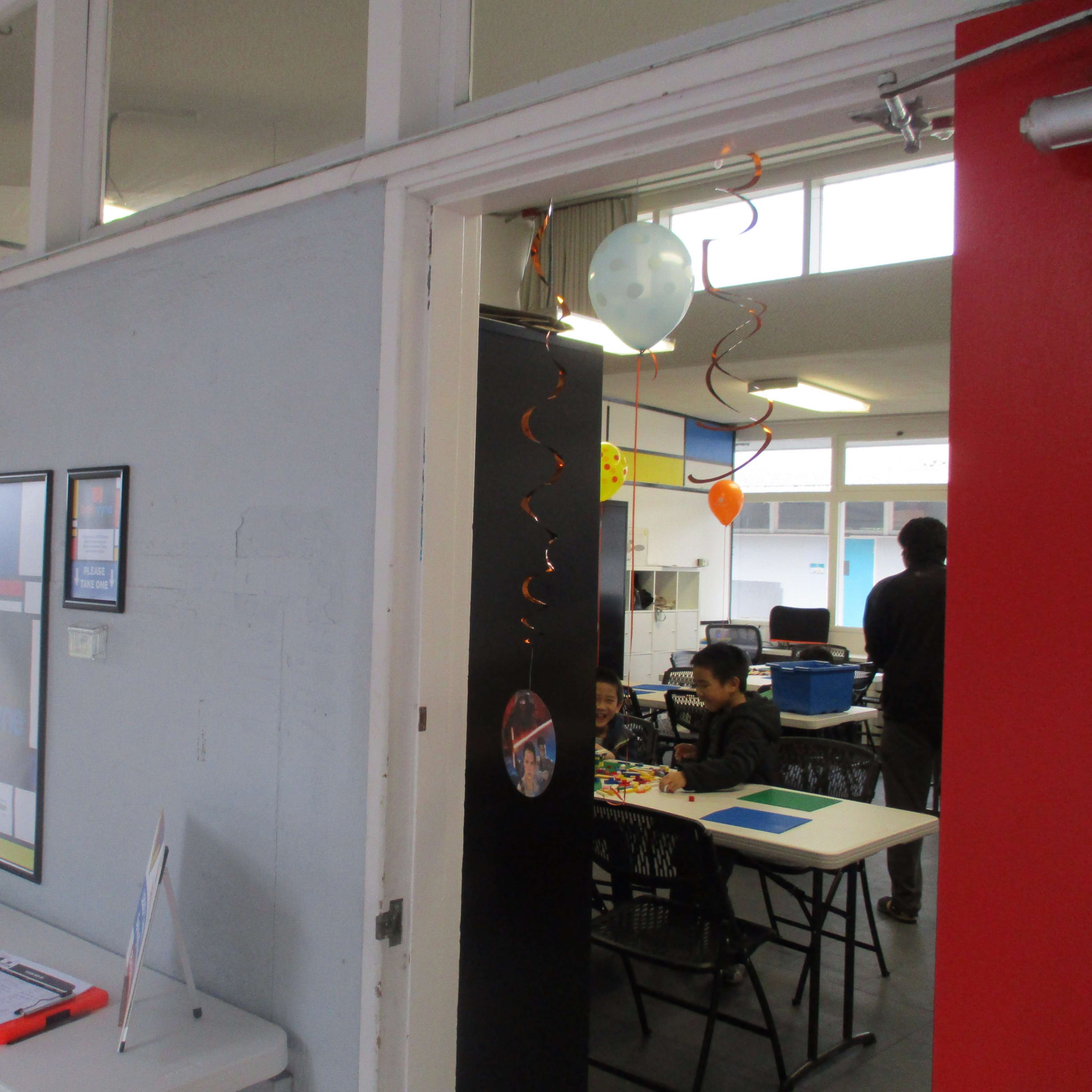 A table is set up in a hallway with a framed poster, while a classroom is visible in the background with a student sitting at a table.