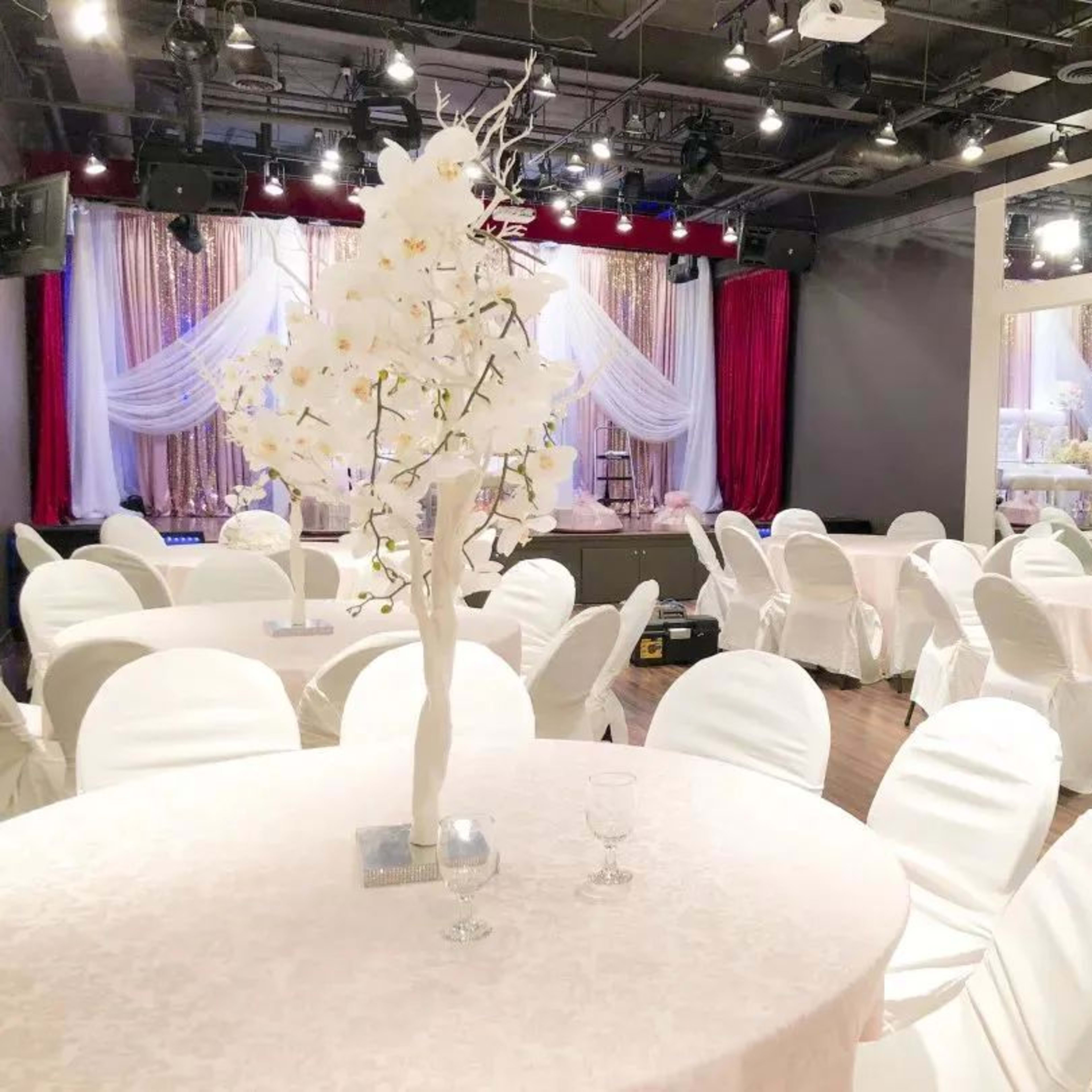 A banquet hall set up with round tables covered in white tablecloths and adorned with white floral centerpieces, accompanied by a stage in the background.