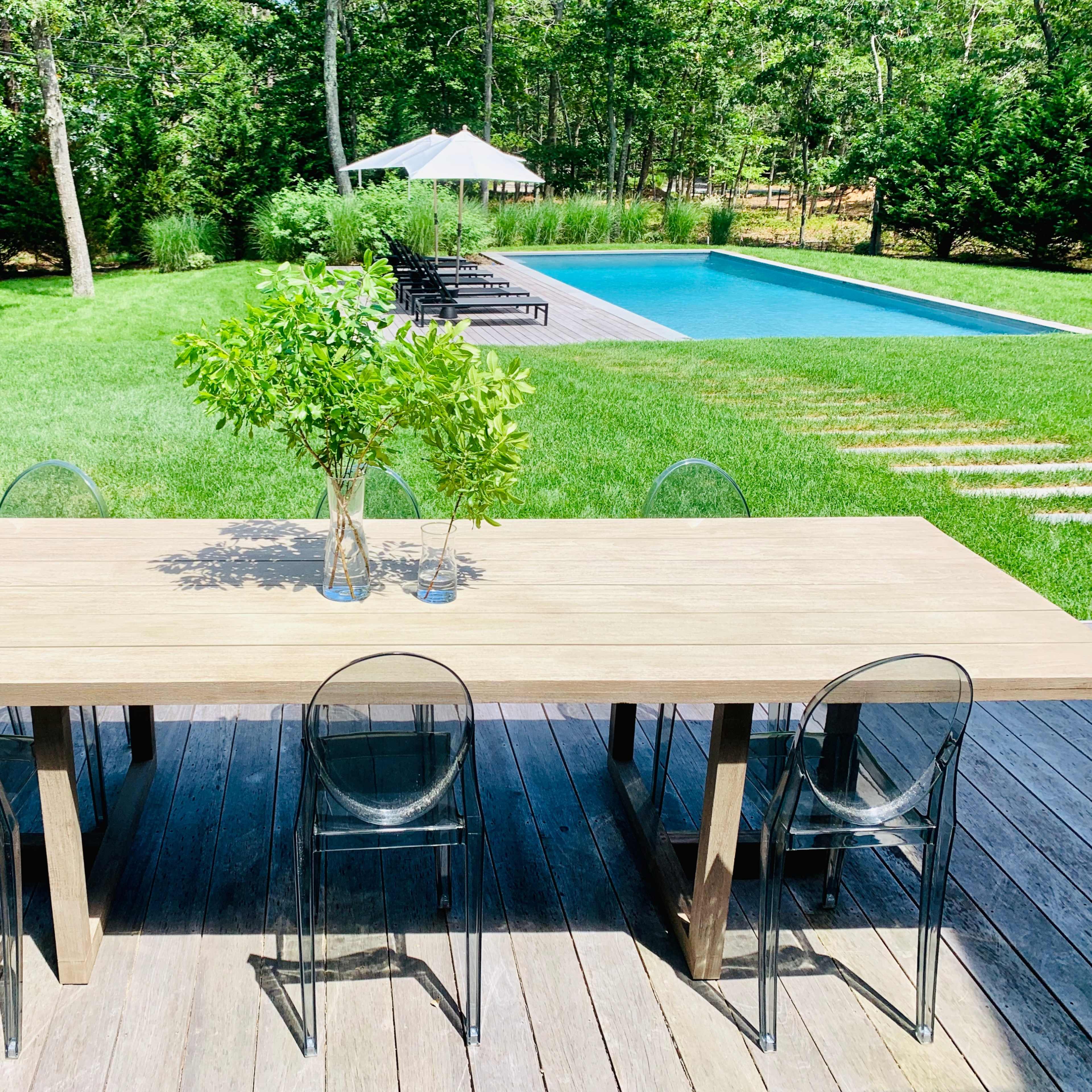 A wooden dining table with transparent chairs is set on a deck, overlooking a lawn with a swimming pool and lounge chairs in the background.