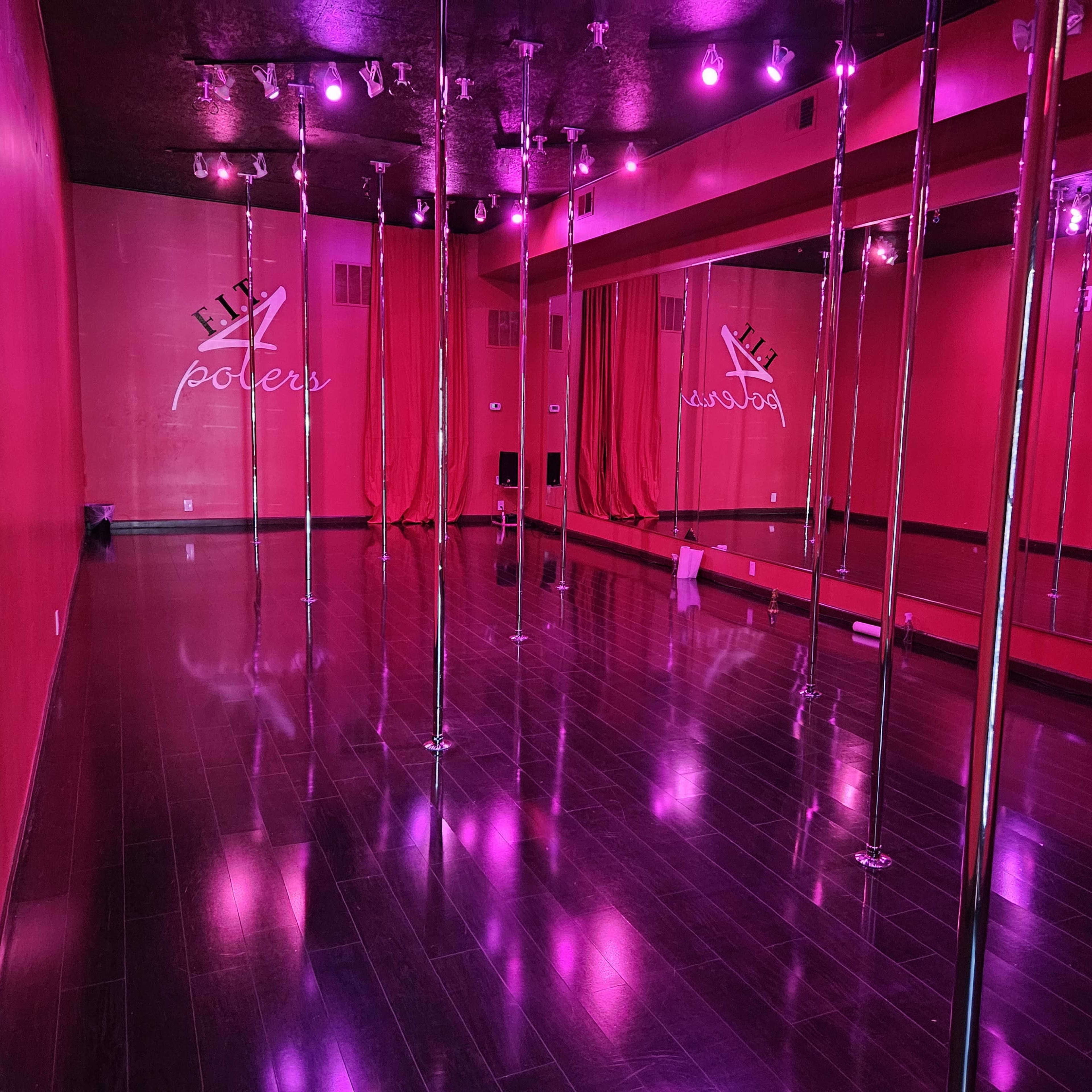 A dance studio with multiple pole dancing poles, mirrored walls, and pink lighting.