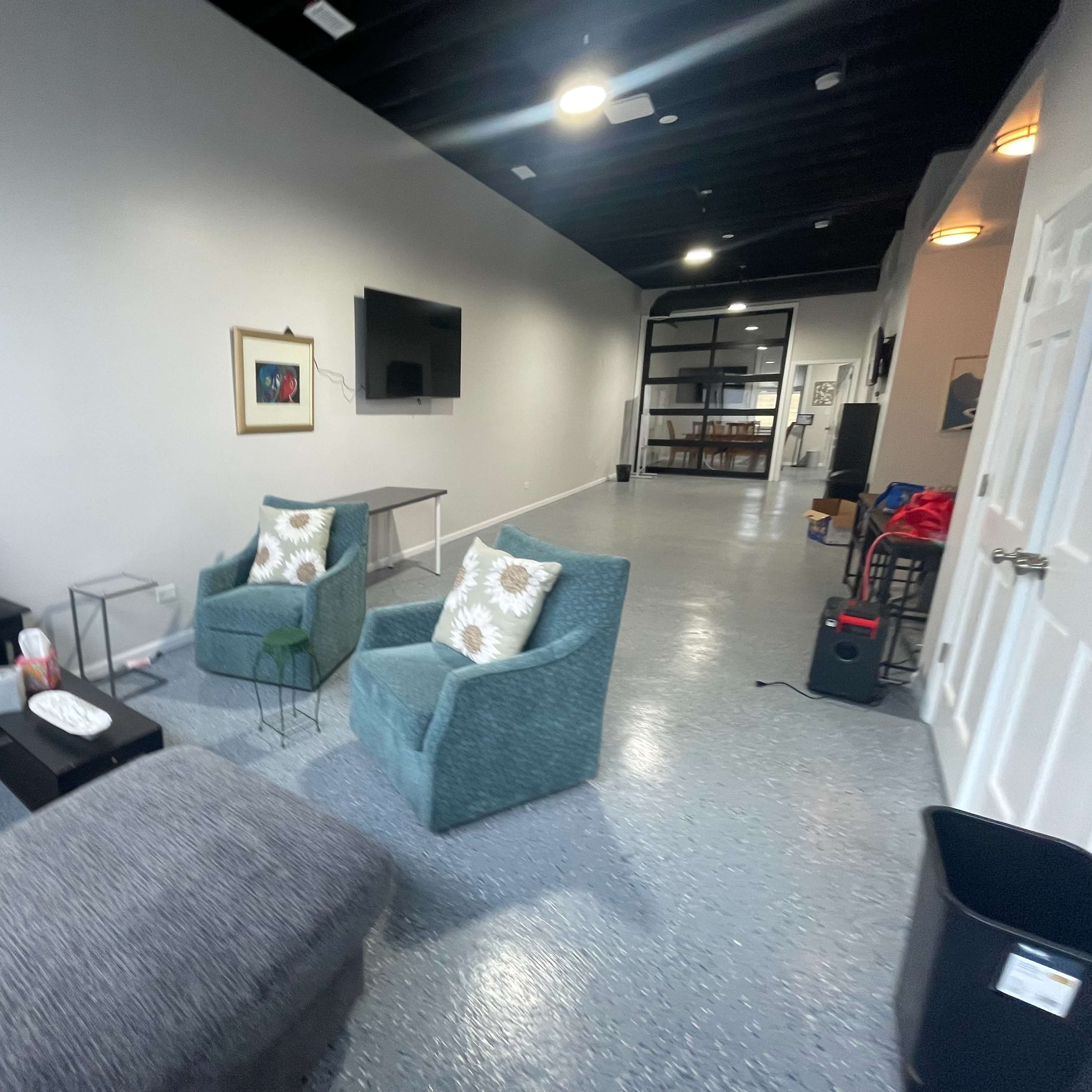 A spacious, well-lit room with a sofa, two chairs, a coffee table, a television mounted on the wall, and a doorway leading to another area.