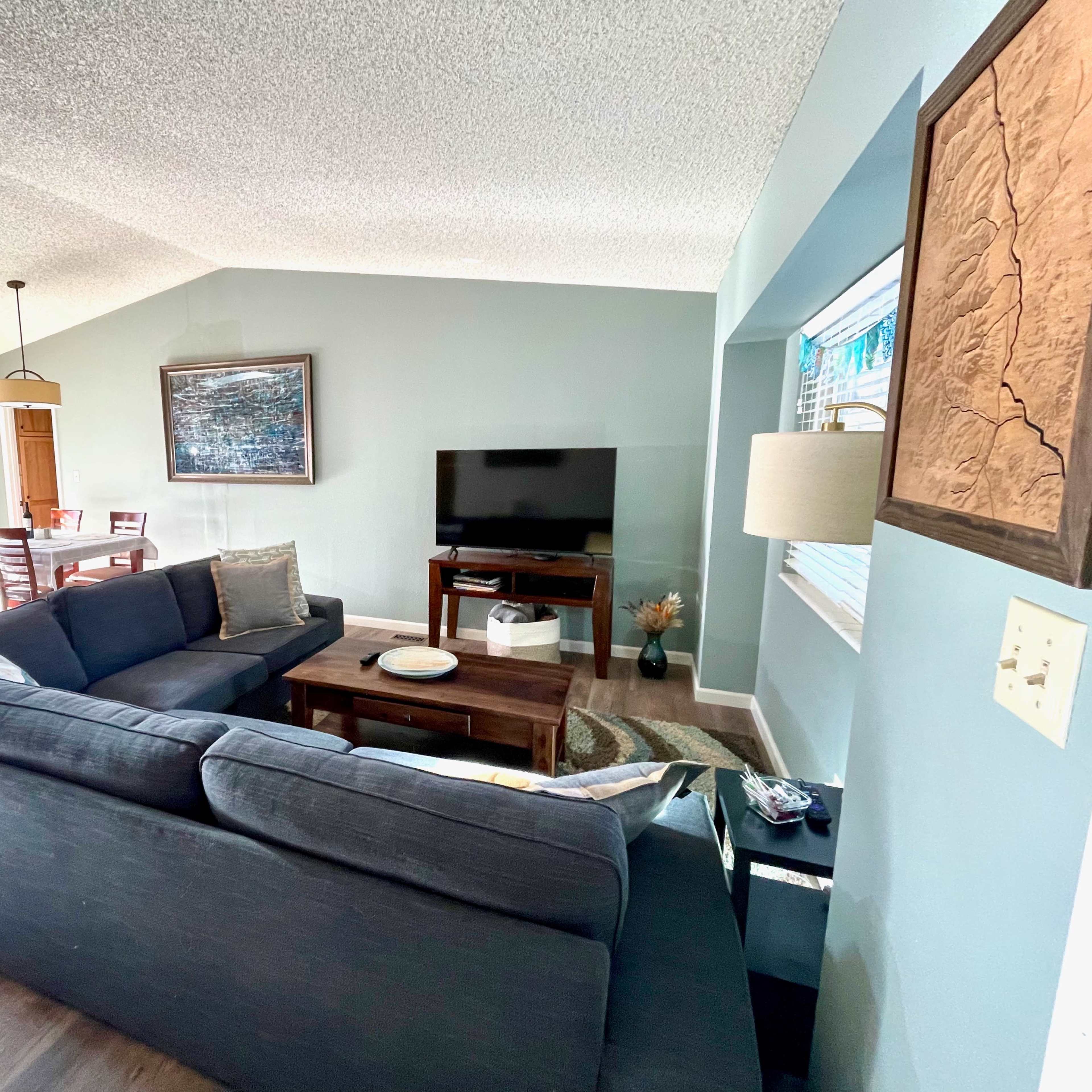 A spacious living room featuring a dark sectional sofa, a coffee table, and a television on a stand, with a dining area and large windows in the background.
