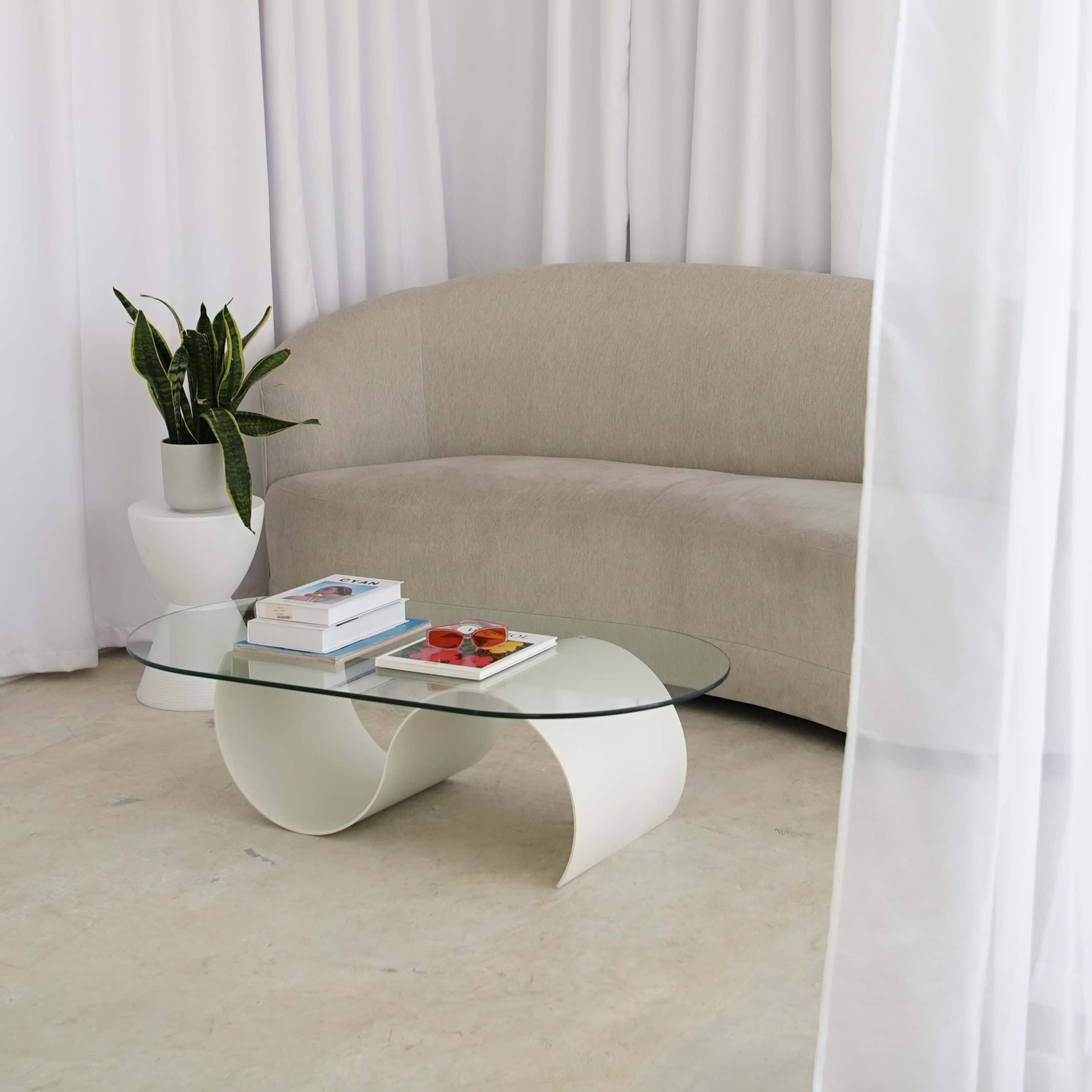 A curved beige sofa and a modern glass coffee table are positioned in front of sheer white curtains and a potted plant.