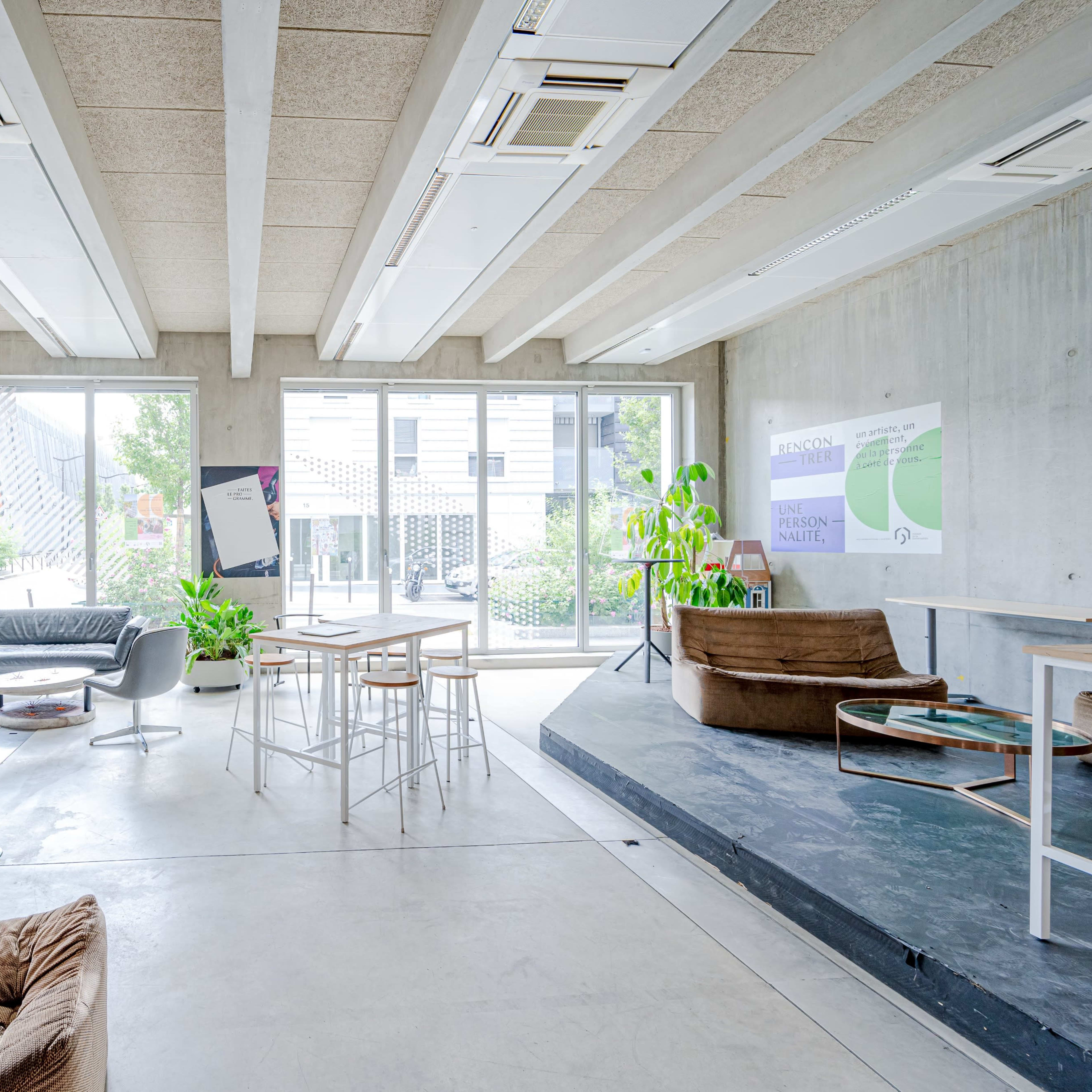 A modern workspace with concrete walls, large windows letting in natural light, and a mix of seating arrangements including chairs, tables, and plants.