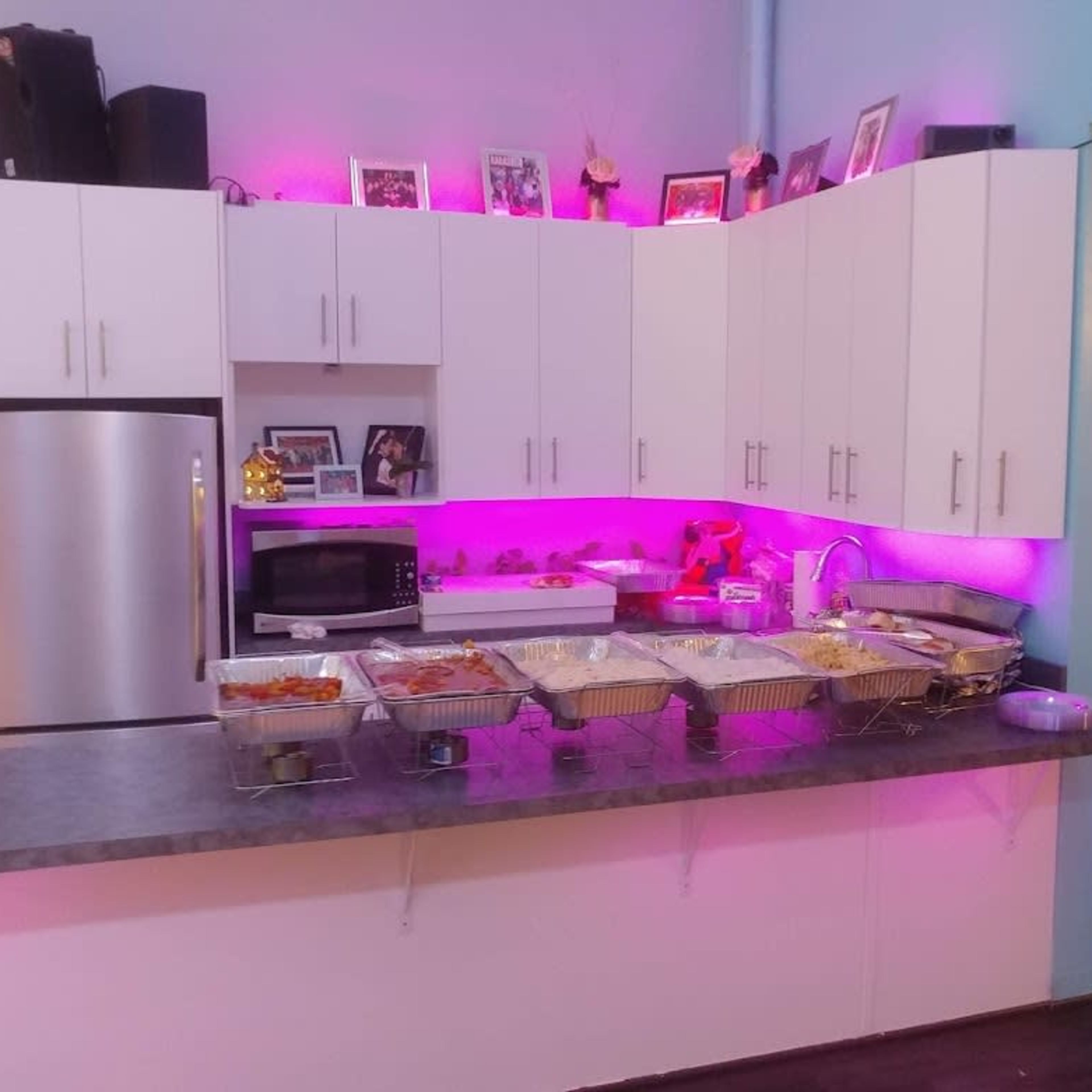 A kitchen counter displays several trays of food under ambient pink lighting.