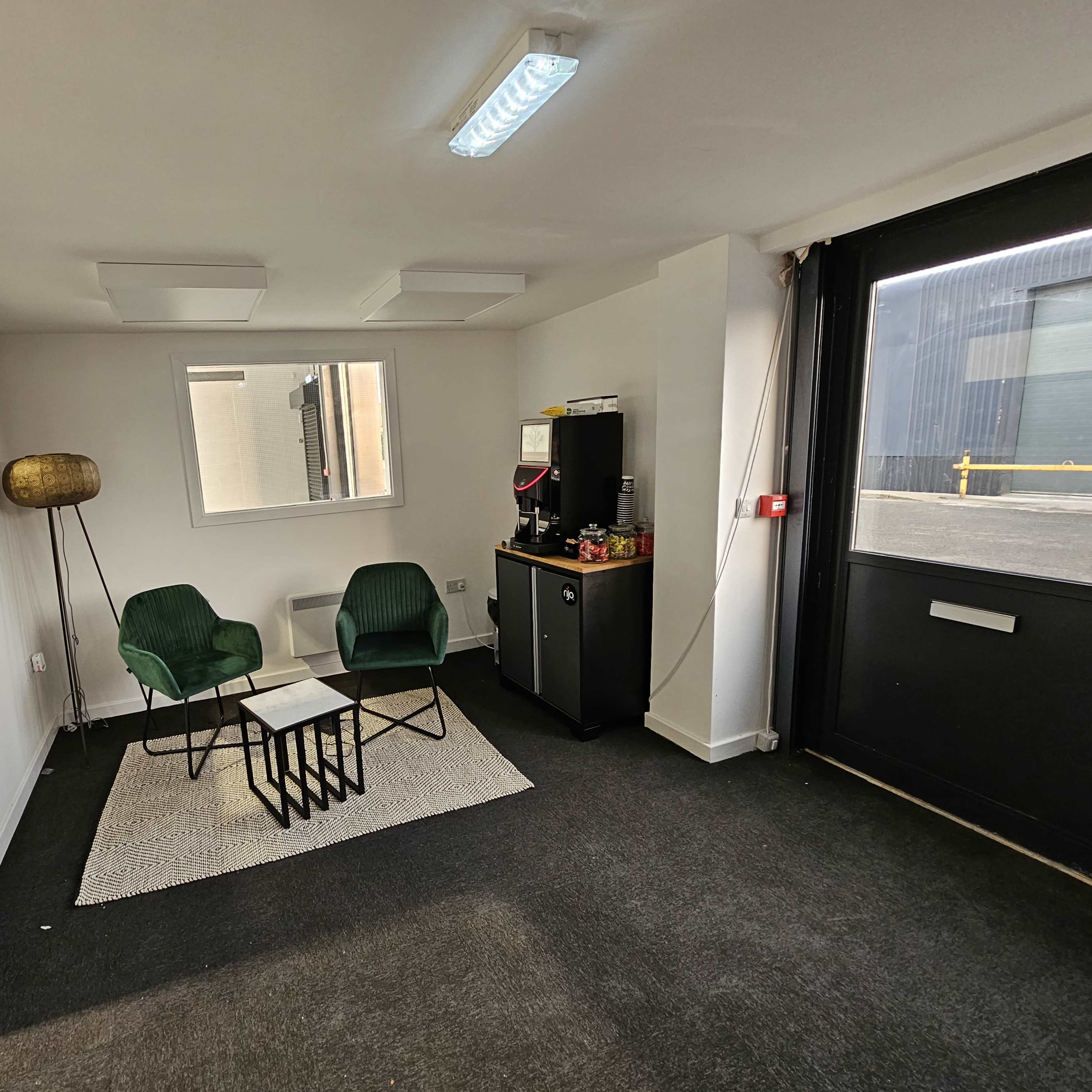 A small office space with two green chairs facing a coffee table on a rug, a refrigerator, and a large glass door leading to an exterior area.