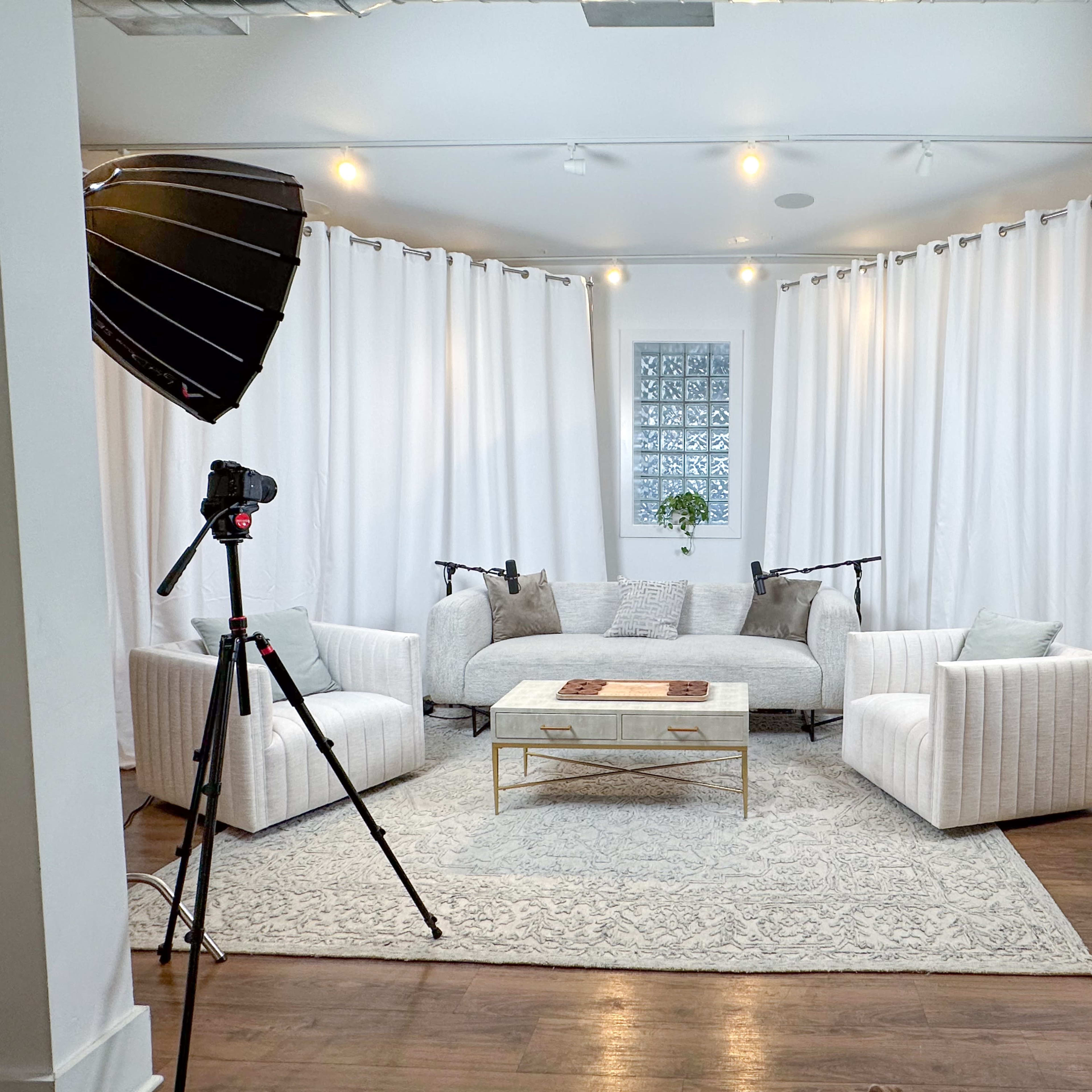 The scene features a bright studio space with a light-colored couch, two armchairs, a coffee table, and soft white curtains, all framed by photography lighting equipment on tripods.