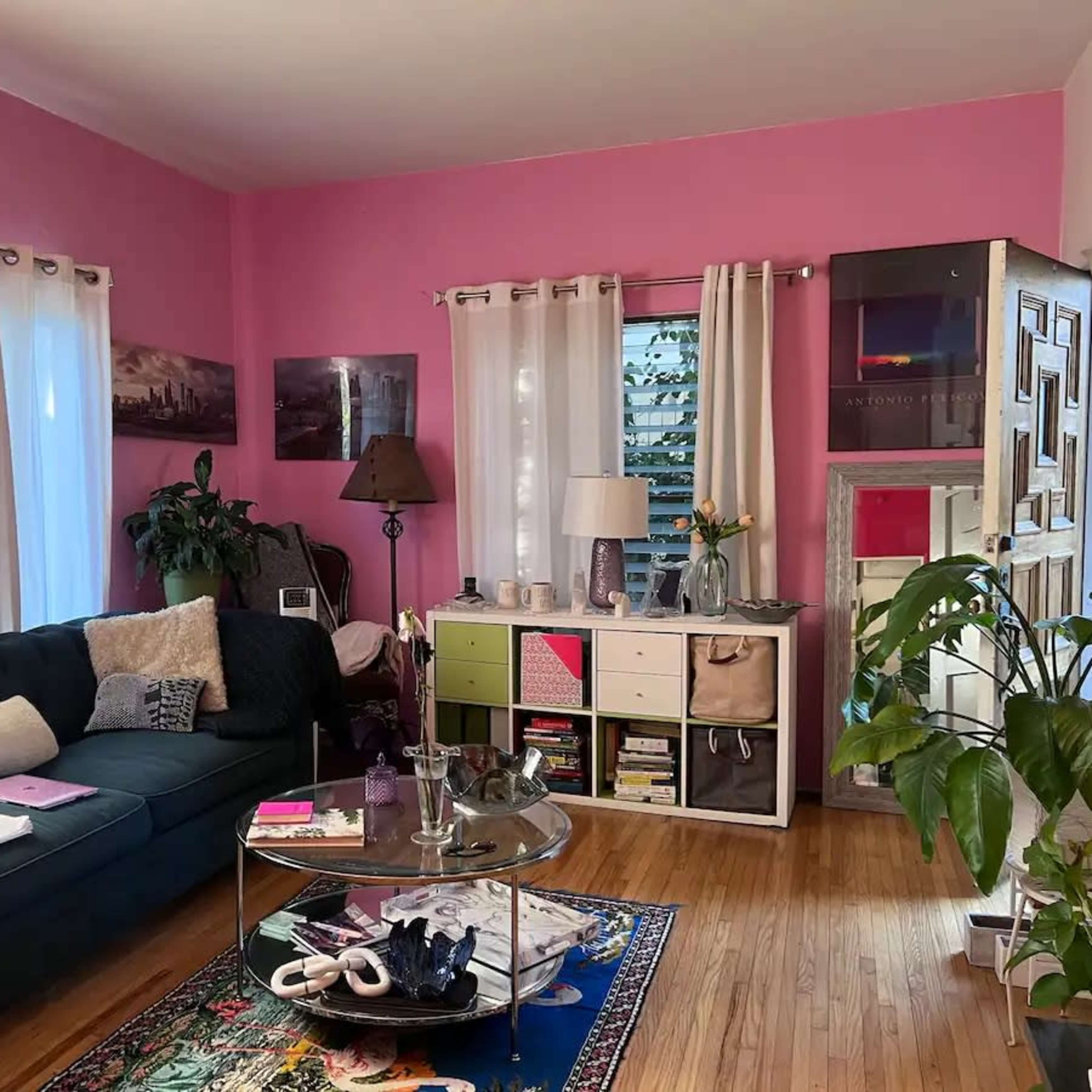 The scene depicts a living room with a pink wall, a blue couch, a glass coffee table, and a mix of plants and decor items.