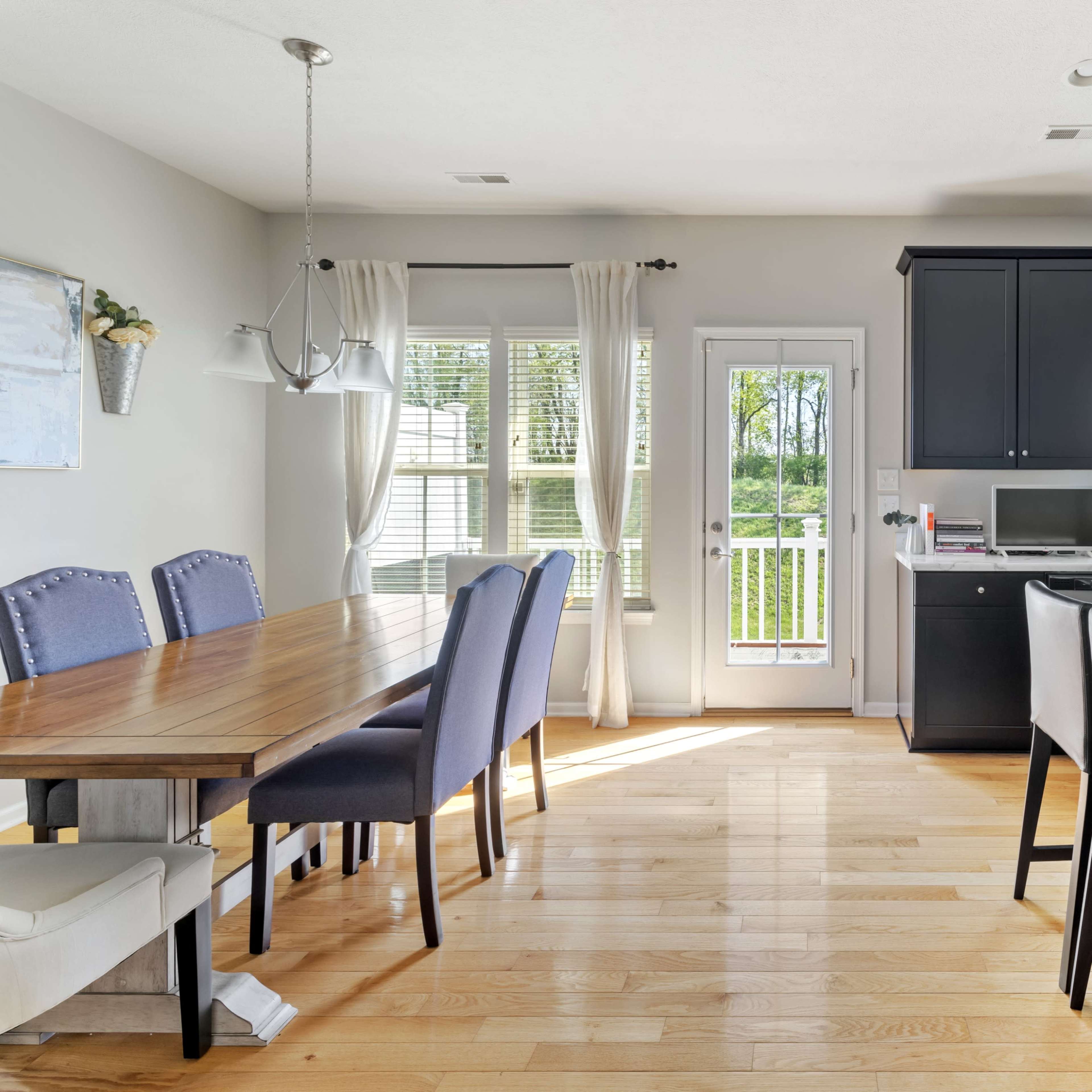 A modern kitchen and dining area features a wooden table with chairs, a kitchen island with bar stools, and large windows overlooking a view.